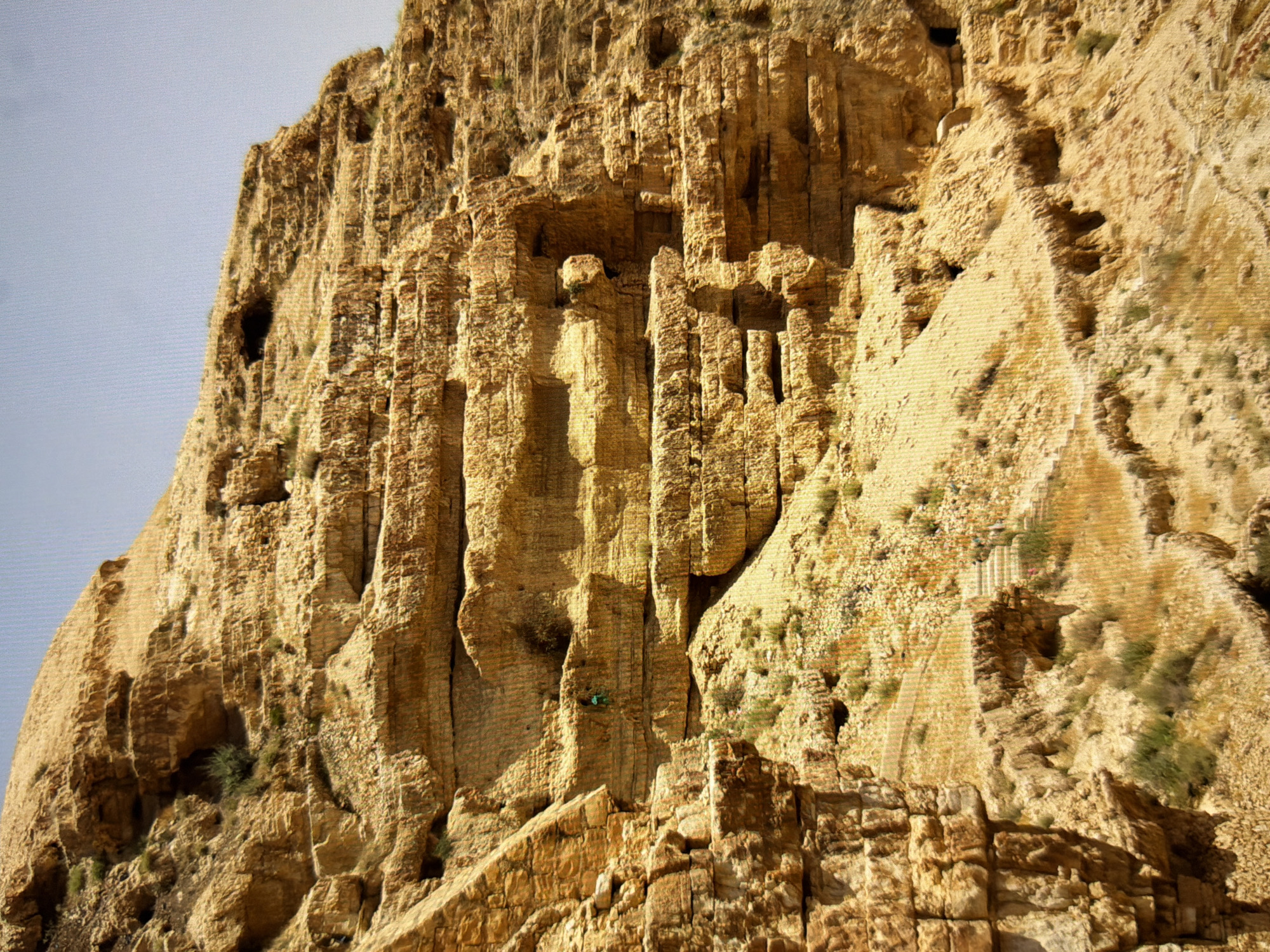 The image displays a rugged, rocky cliff face, characterized by layered sedimentary rock in shades of tan and yellow. Sparse green vegetation grows in crevices and on ledges. A narrow, winding path is visible traversing the lower portion of the cliff, with what appear to be steps carved into the rock in some areas. In the middle section of the cliff, a small, dark opening suggests a cave or alcove. A person, wearing a green garment, is visible on a ledge, seemingly engaged in climbing or exploring the rock face. The sky above is a pale, hazy blue. The scene depicts a natural, remote environment. There is no indication or visual evidence within the image to suggest this location is in Kyiv, Ukraine.