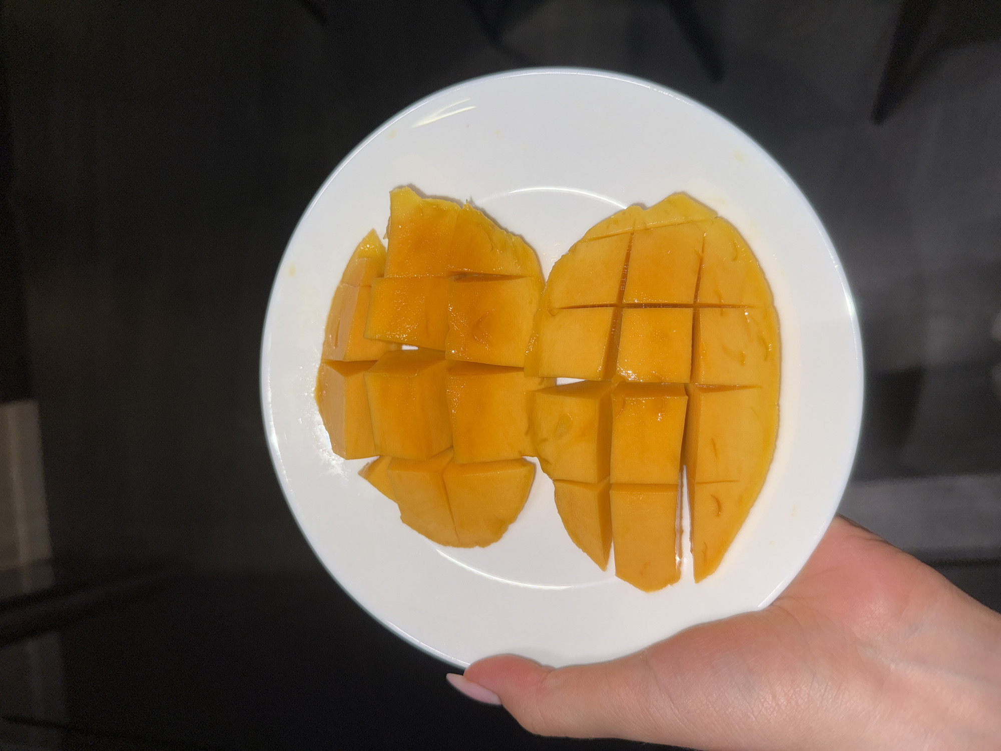 A plate with two mango halves, cut into a grid pattern, is held in a person's hand. The plate is white and round. The mango flesh is a bright yellow-orange color. The hand is fair-skinned with pink nail polish. The background is a dark, indistinct color. The location is Unknown, Unknown.