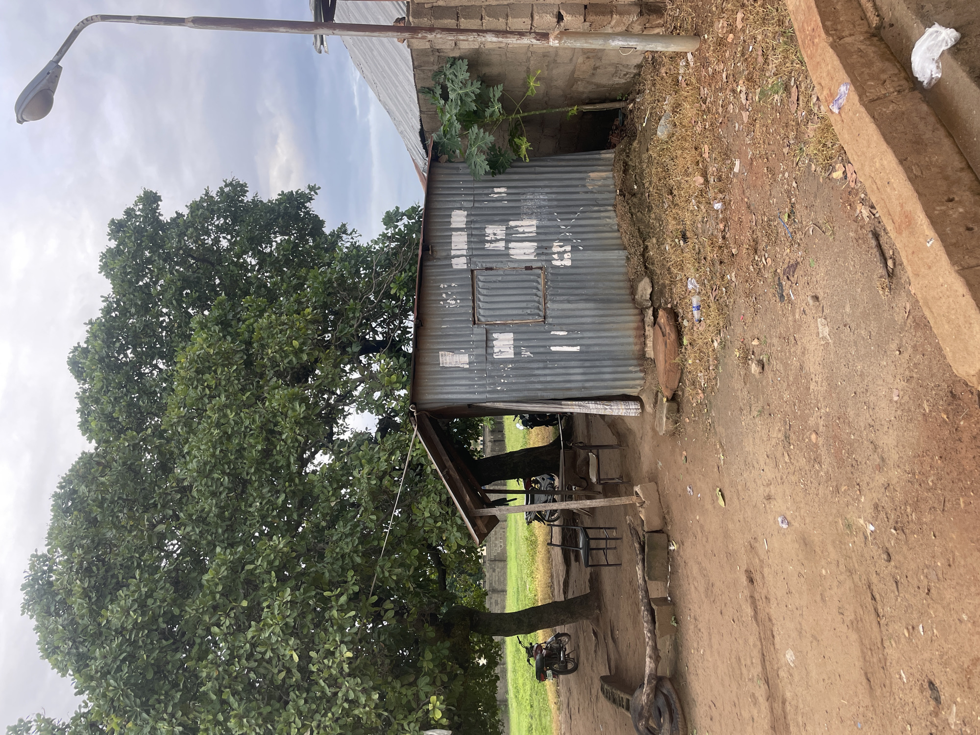 The image depicts an outdoor scene in Keffi, Nigeria, featuring a small, weathered corrugated metal structure with a wooden roof. The structure appears to be a makeshift shop or shelter, with a window covered by a metal grate. In front of it, two chairs and a table are set up on a dirt ground. A motorbike is parked to the left of the structure, near a large tree. Another motorbike is visible further back, partly obscured by the trees. A tall streetlight stands to the right of the metal building, next to a concrete wall. The ground is covered in dirt, dry grass, and scattered debris, including plastic bottles and bags. In the background, a grassy field is bordered by a low brick wall, and additional structures with corrugated metal roofs are partially visible. The sky is overcast.