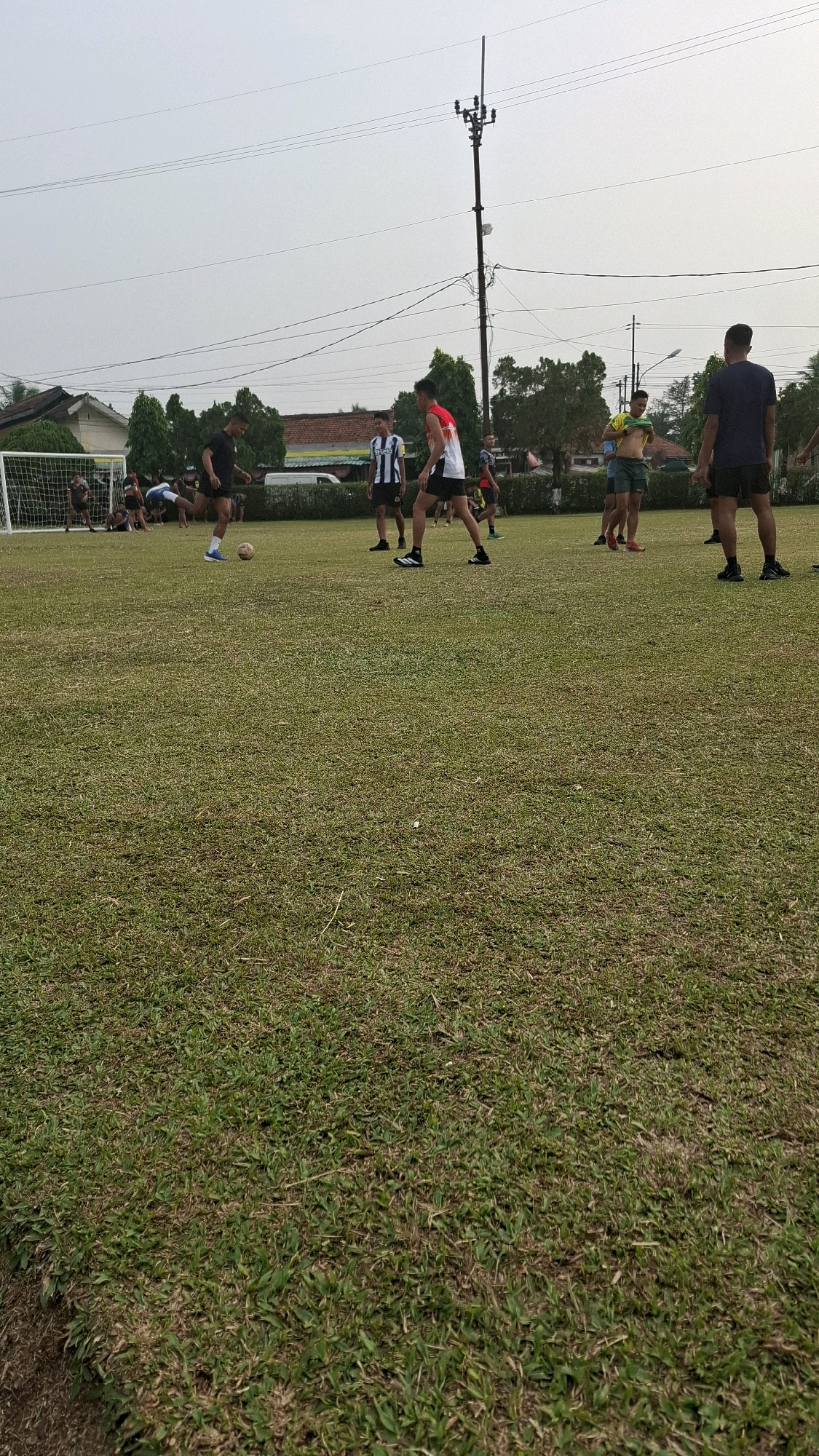 In Sukaraja, Indonesia, a group of young men are engaged in a soccer practice on a grassy field. The field is marked with a soccer goal at the left side of the frame. Several individuals are actively participating in the game, with one person in the foreground kicking a soccer ball. Other individuals are positioned around the field, either observing or preparing to join the game. The background reveals residential buildings and trees, suggesting an urban or suburban setting. A utility pole with wires stands prominently in the middle ground. The sky is overcast, indicating a cloudy day.