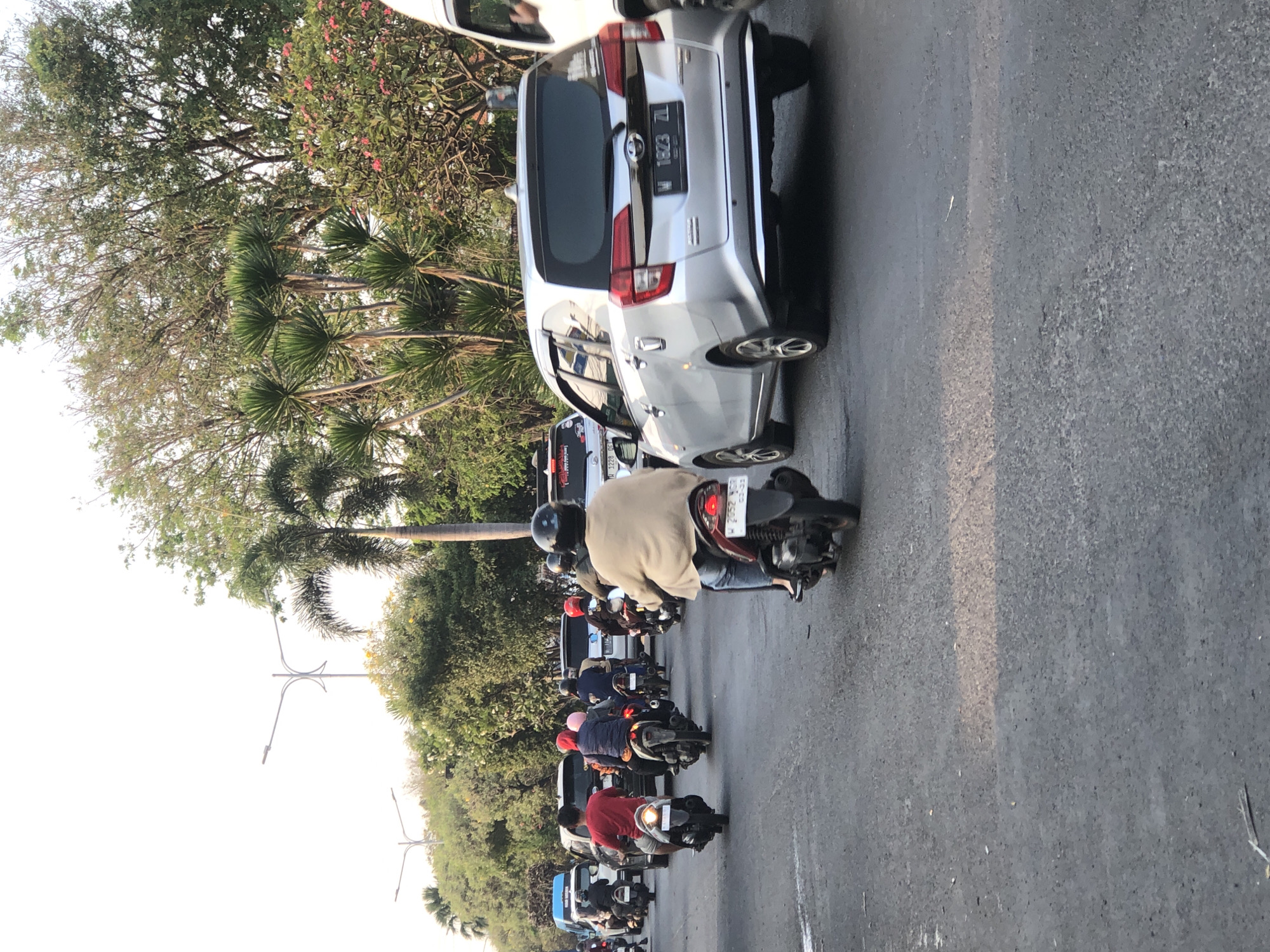 The image shows a street scene in Ngaban, Indonesia, with multiple vehicles and people. The road is paved and appears to be a multi-lane street. On the right side of the image, a silver car with license plate "W 1823 ZL" is visible, and partially obscured by a white vehicle. In the center of the image, several motorcycles are in motion. One motorcycle in the foreground has a rider wearing a beige jacket and jeans, and a black helmet. Behind this motorcycle, another motorcycle carries two people, one in a red shirt and the other in a pink headscarf. Further back, other motorcycles and cars are visible in the traffic. Trees and palm trees line the sides of the street, with some flowering bushes in the background. Streetlights are also present, indicating an urban or suburban setting. The sky is bright, suggesting daytime, likely late afternoon or early morning given the lighting.