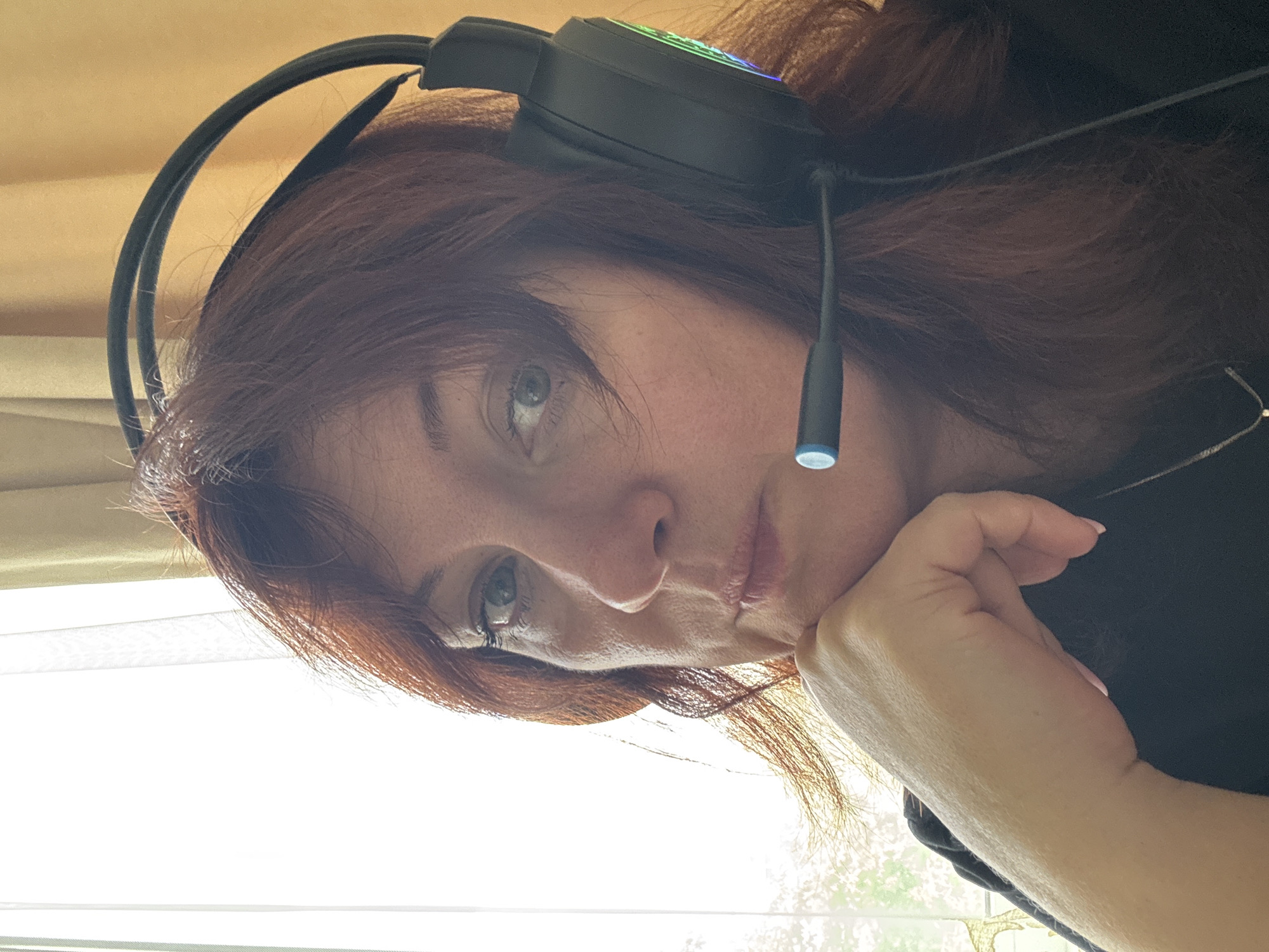 A woman with auburn hair is looking into the camera with a neutral expression, resting her chin on her hand. She is wearing a black headset with a microphone. The background consists of a window with sheer white curtains and yellow drapes. The image appears to be taken indoors, with natural light coming from the window. There is no discernible information in the image that places it in Bila Tserkva, Ukraine.