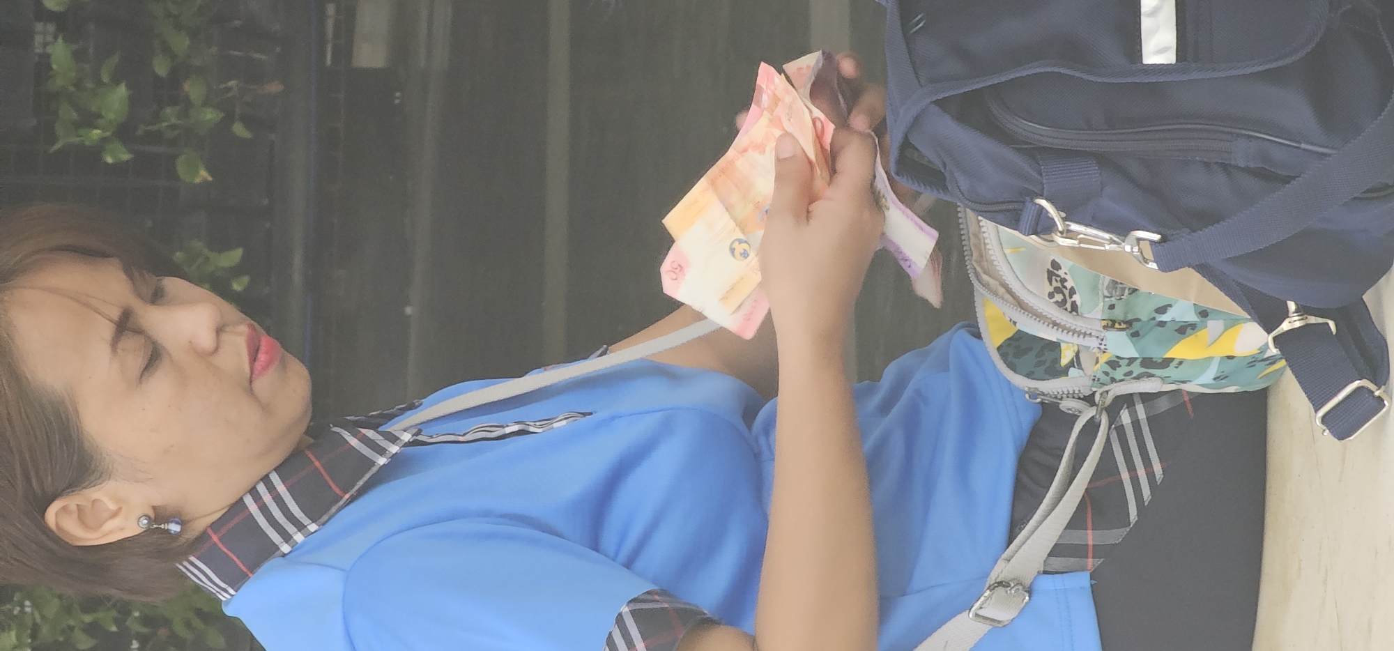 In Makati City, Philippines, a woman with short brown hair, wearing a blue collared shirt with a plaid trim, is holding several Philippine peso bills, appearing to be counting them. She has on lipstick and a dangling earring. She is seated next to a patterned bag and a dark blue bag. The background is out of focus and appears to be an outdoor setting with greenery.