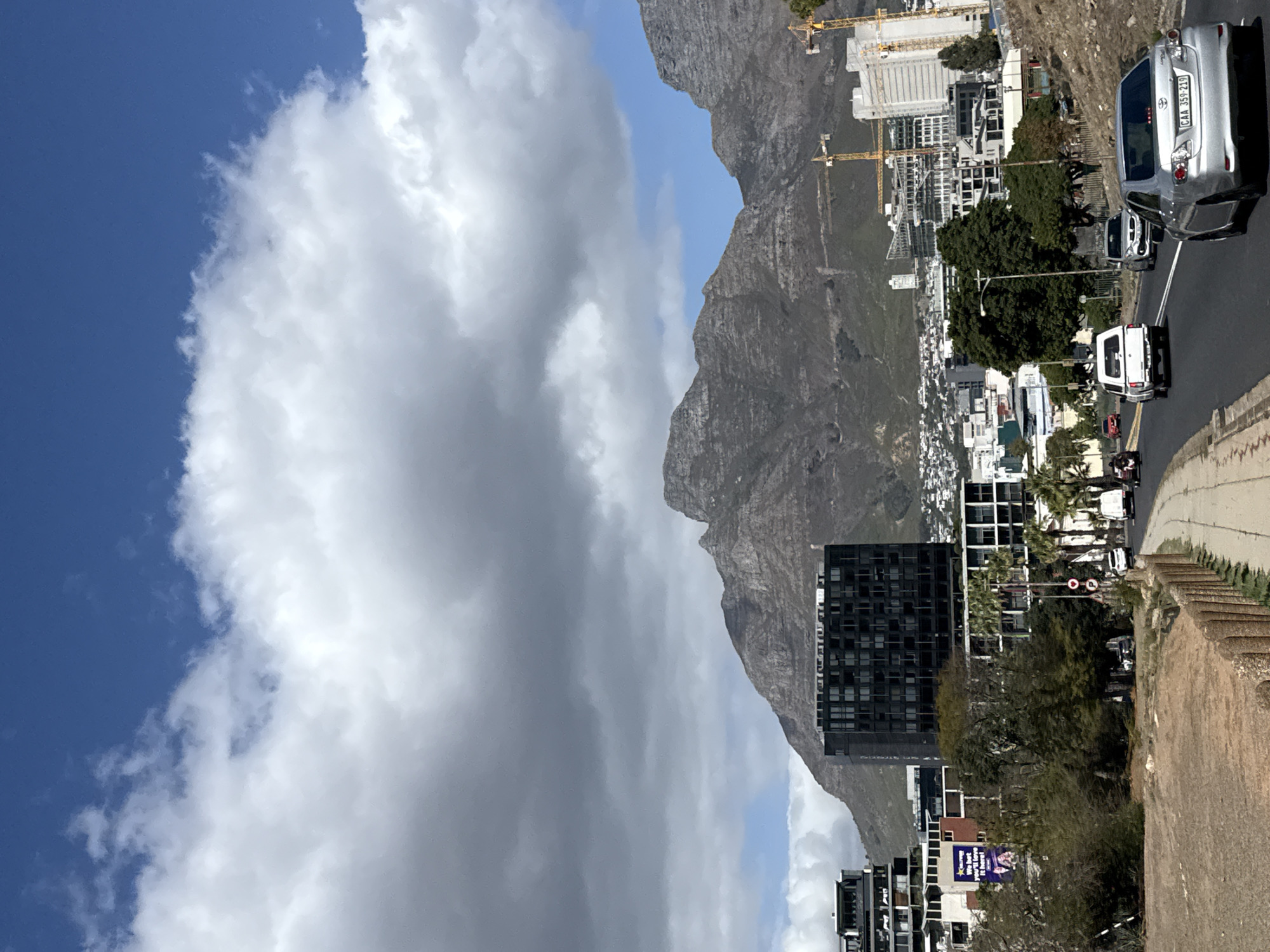 The image depicts a view in Cape Town, South Africa, featuring Table Mountain in the background. A mix of buildings, including modern high-rises and smaller structures, are situated along the mountain's lower slopes. Construction cranes are visible among the buildings. A road with vehicular traffic runs along the edge of the scene, including cars and what appears to be a motor scooter. The foreground shows a wall or embankment. The sky is partially clear with blue patches interspersed with large, billowing clouds.