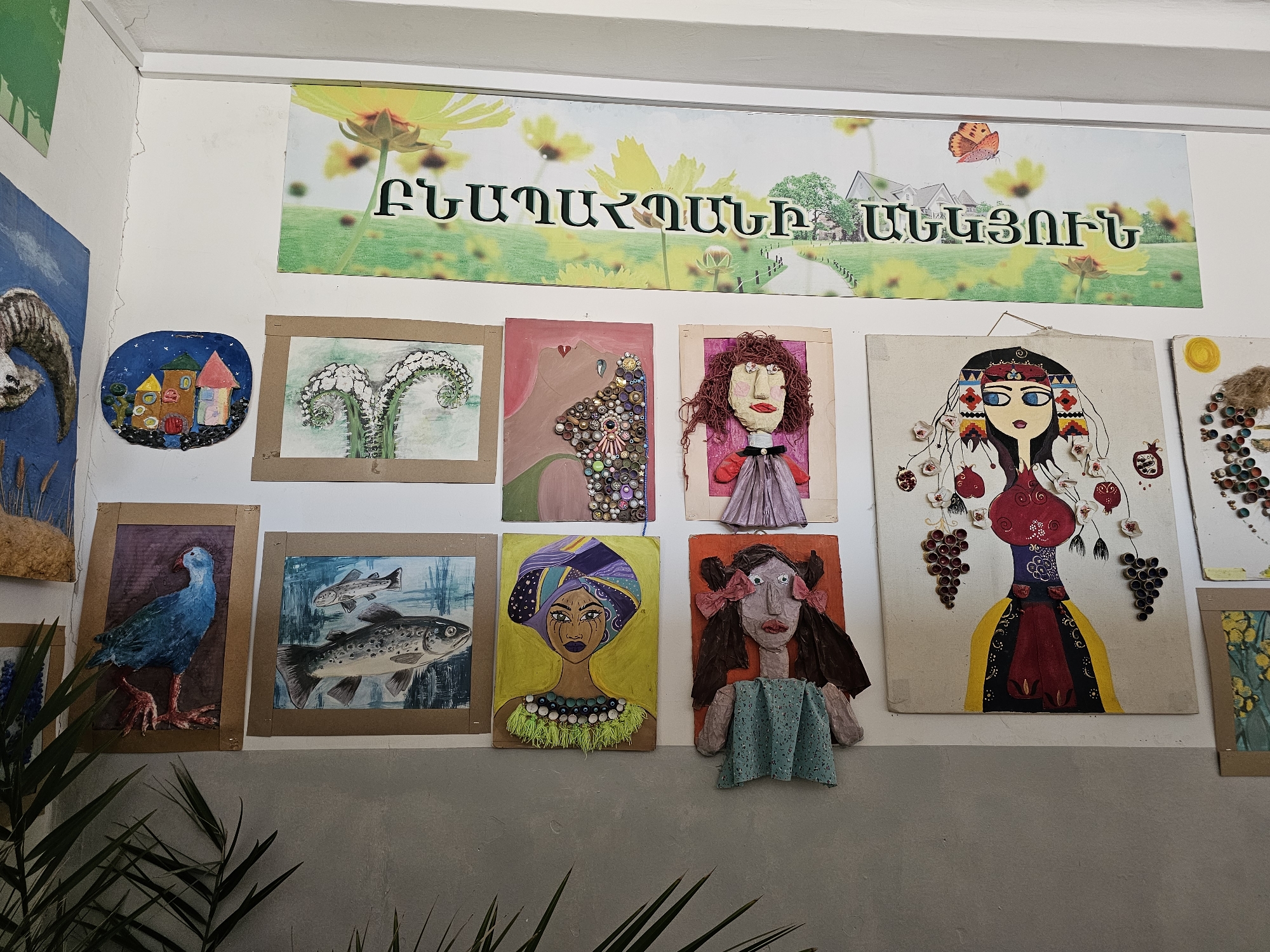 The image shows a collection of children's artwork displayed on a white wall in what appears to be a school or community center. The artwork includes a variety of mediums and subjects, such as paintings of animals (fish and a bird), landscapes (house scene), and portraits of people using mixed media. A banner in Armenian text is visible above the artwork. The location is likely in Yerevan, Armenia, based on the language of the banner. The scene lacks people. A potted plant is visible in the lower left foreground. The wall is two-toned; with white on top and grey on the bottom.