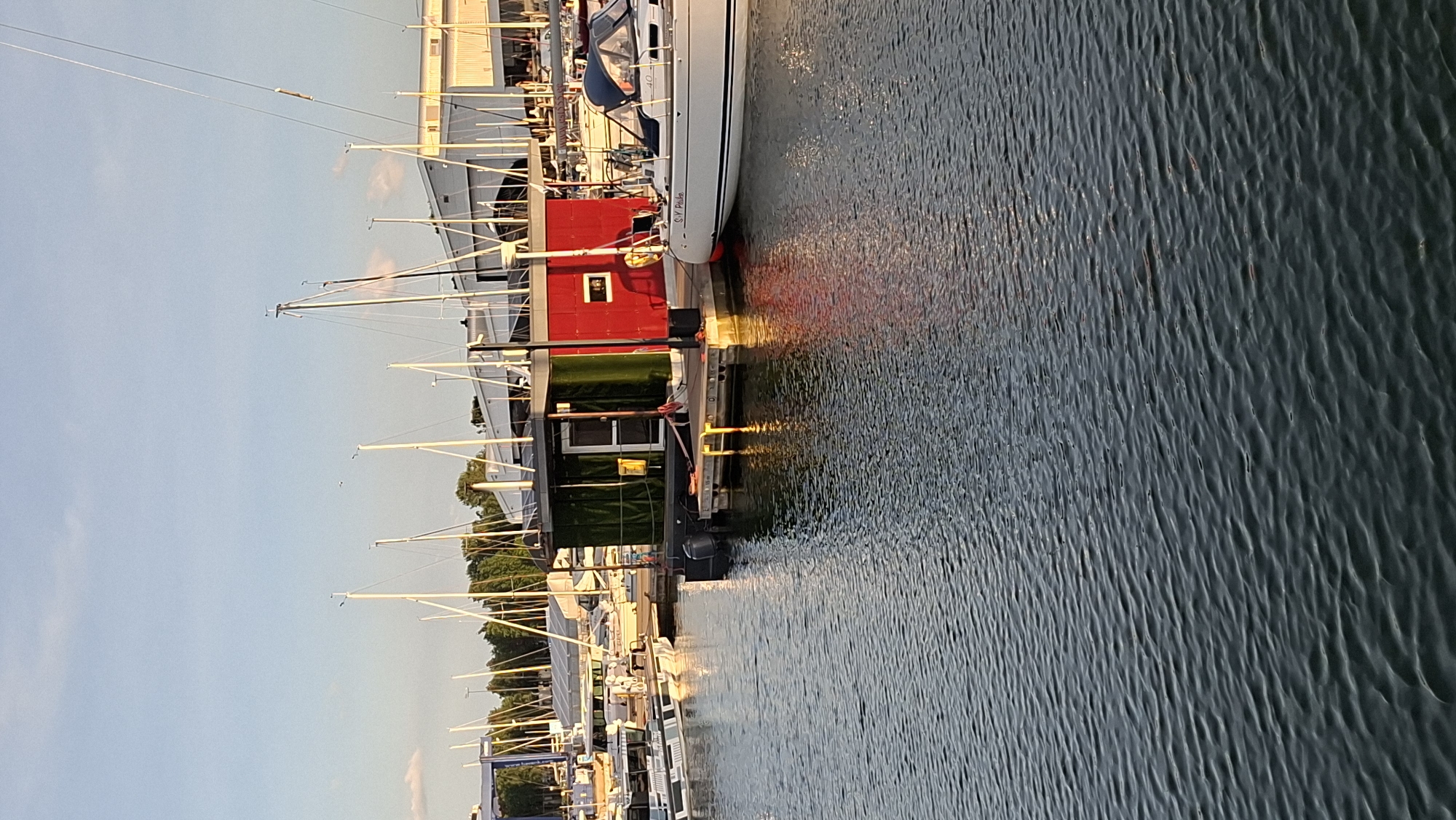 An outdoor, medium shot captures a marina on a clear day with calm water. In the foreground, the rippling water reflects the warm glow of the setting sun, casting colorful hues across its surface. Moored boats of various sizes line the dock, with their masts creating a striking silhouette against the sky. A houseboat, painted red and green, sits prominently in the middle ground, its simple structure contrasting with the more elaborate sailboats. In the background, a row of buildings with large windows and slanted roofs suggests a commercial area or boathouse. The overall impression is one of tranquility and leisure, with the marina bathed in the soft light of early evening.