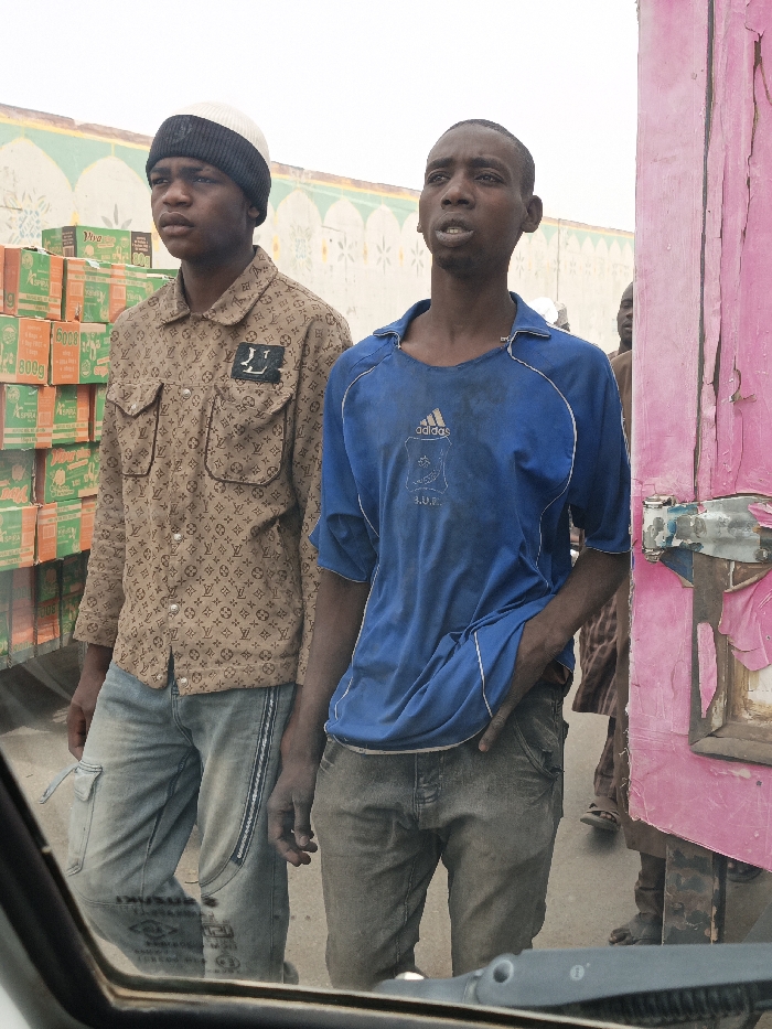 In Fagge C, Nigeria, two young males are observed standing outdoors, viewed from within a vehicle. The male on the left, wearing a black and white knit beanie, a tan long-sleeved shirt with a repeating monogram pattern and an 'LV' logo patch, and light blue denim jeans, stands with a neutral expression, looking slightly to his left. Behind him and to his left, stacks of green and orange boxes labeled "VIVA," "ESPRERA," "600g," and "800g" are visible, possibly on a transport vehicle. The male on the right, wearing a dirty blue short-sleeved t-shirt with an "adidas" logo and dark denim jeans, stands with his left hand in his pocket, looking slightly upward and to his right, his mouth slightly open. To his immediate right, a peeling, faded pink wooden or metal structure with a visible metal latch mechanism occupies the right edge of the frame. In the background, behind both individuals, a light-colored wall with decorative green and white arched and geometric patterns is visible. The ground appears to be an unpaved or dirt surface. Other partial figures are dimly visible in the background behind the right subject. The sky is bright and overcast.
