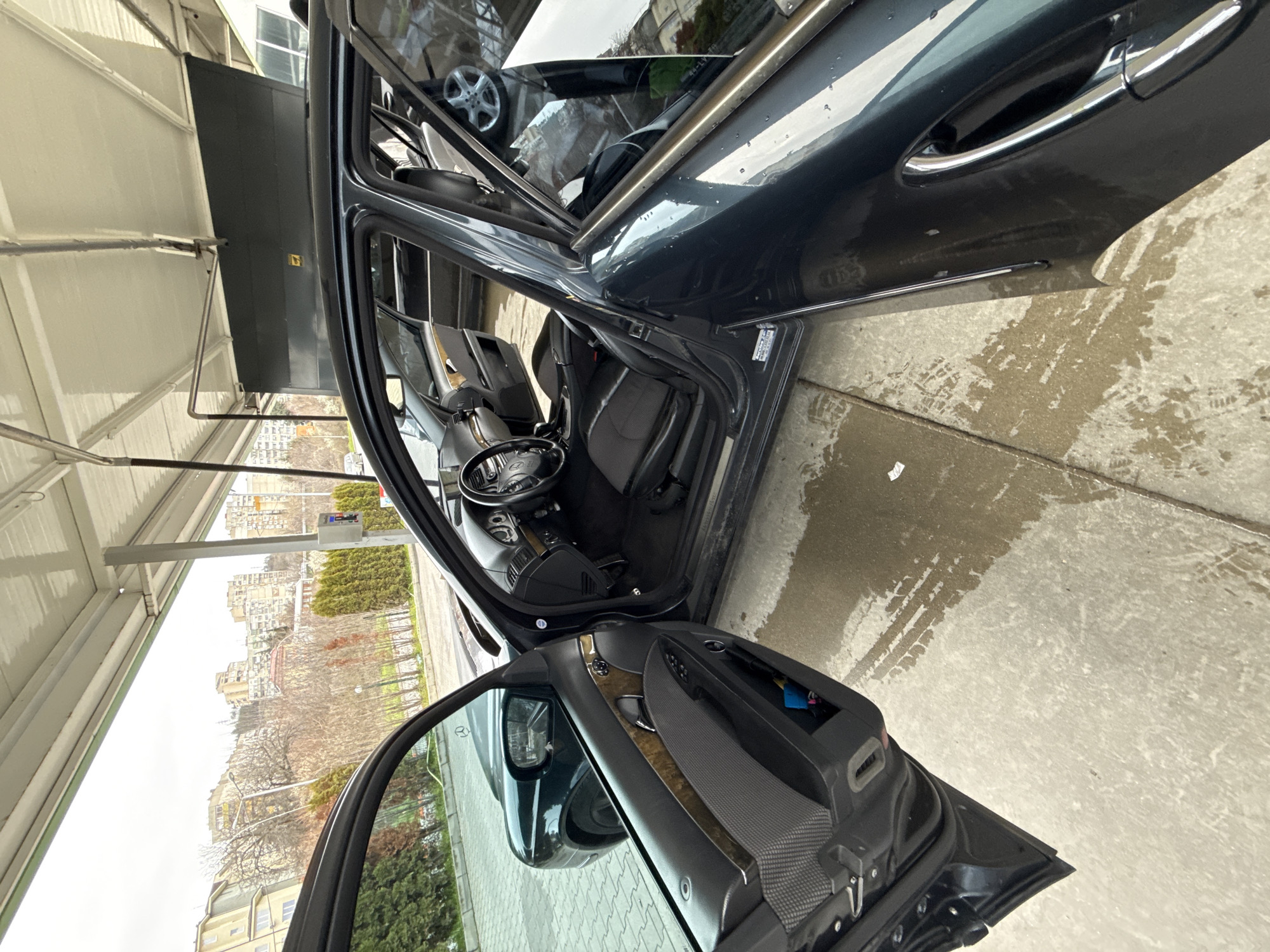 The image shows the interior of a dark-colored car with open doors, parked under a structure resembling a gas station canopy. The car's interior is visible, including the steering wheel, seats, and dashboard. The exterior background features buildings and trees, typical of an urban setting.
Based on visual clues such as architectural style, vegetation, and potential street signs, it is likely that the location of this scene is somewhere in the city of Stara Zagora, Bulgaria. However, without clear visible landmarks or street signs, pinpointing the exact location is not possible.