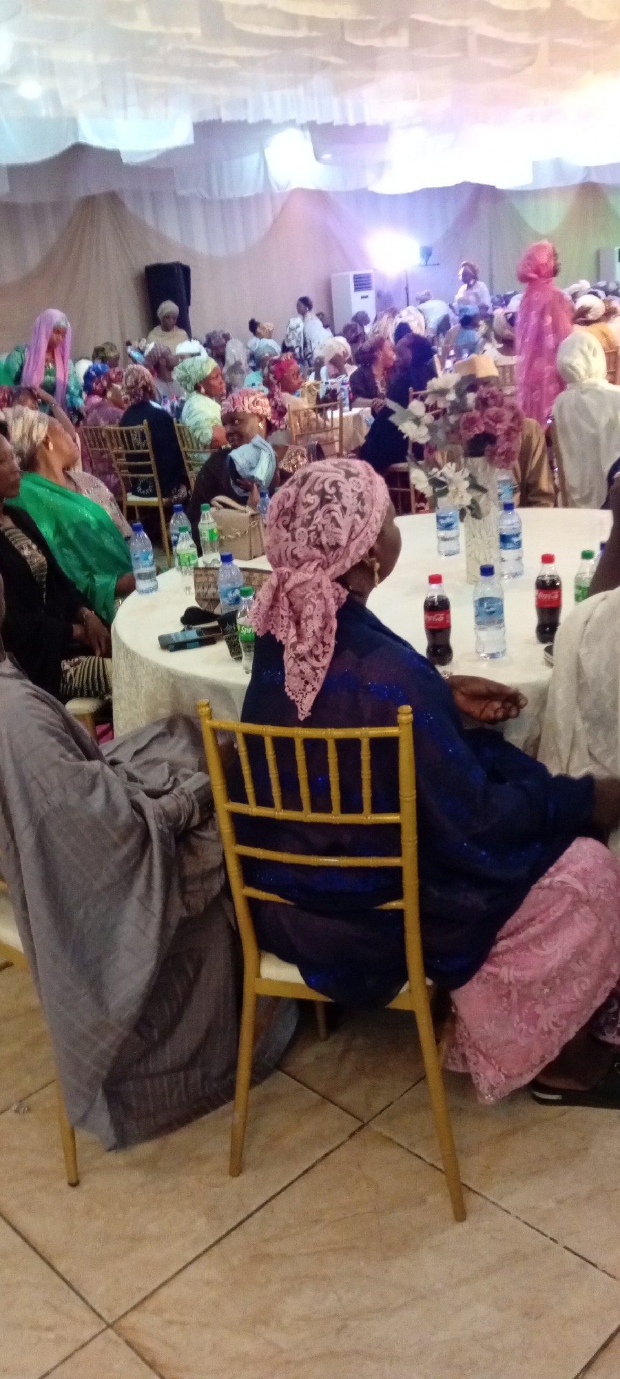In Gandu Albasa, Nigeria, a formal gathering is taking place, likely a social event or celebration, within a large indoor space adorned with draped white fabric creating a tent-like ceiling. The room is filled with many individuals, predominantly women, dressed in vibrant traditional attire and head coverings. Several round tables are set up, covered with white tablecloths and decorated with floral arrangements in vases. Bottles of water and soda, along with mobile phones, are visible on the tables, suggesting a seated dining or reception setting. The floor is tiled with large, light-colored square tiles. The lighting appears to be a mix of ambient and spotlighting, creating a warm, illuminated atmosphere. The focus is on the people engaged in conversation and enjoying the event, showcasing a community coming together.
