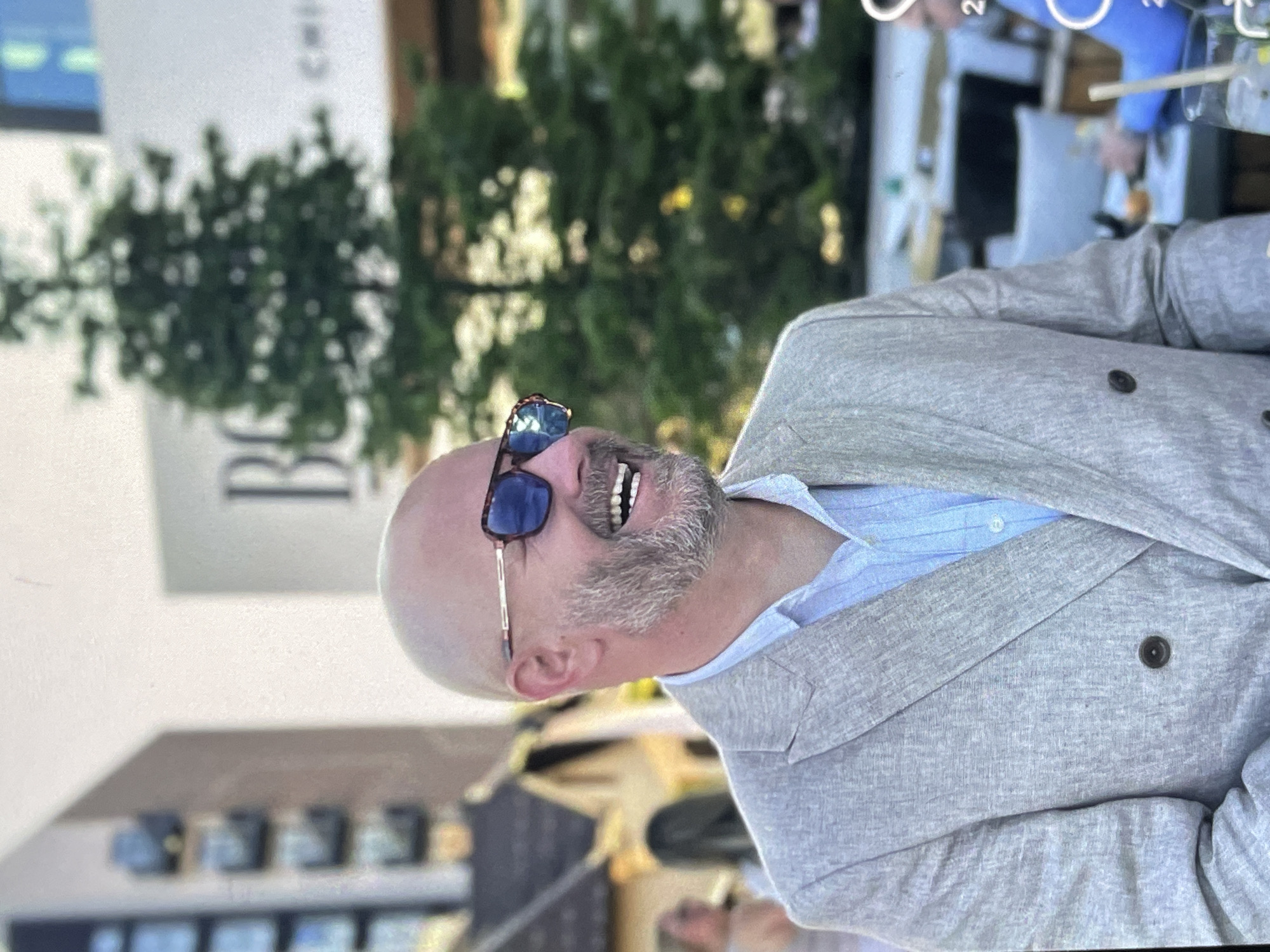 The image shows a bald, Caucasian man with a beard, wearing sunglasses, a light blue shirt, and a light-colored double-breasted blazer. He is smiling broadly. The man is in an outdoor setting, possibly a restaurant or event. In the background, there is a tall, green tree and a building with "BG" logo visible. There are other people in the background, some seated at tables. The overall scene appears to be a social gathering.