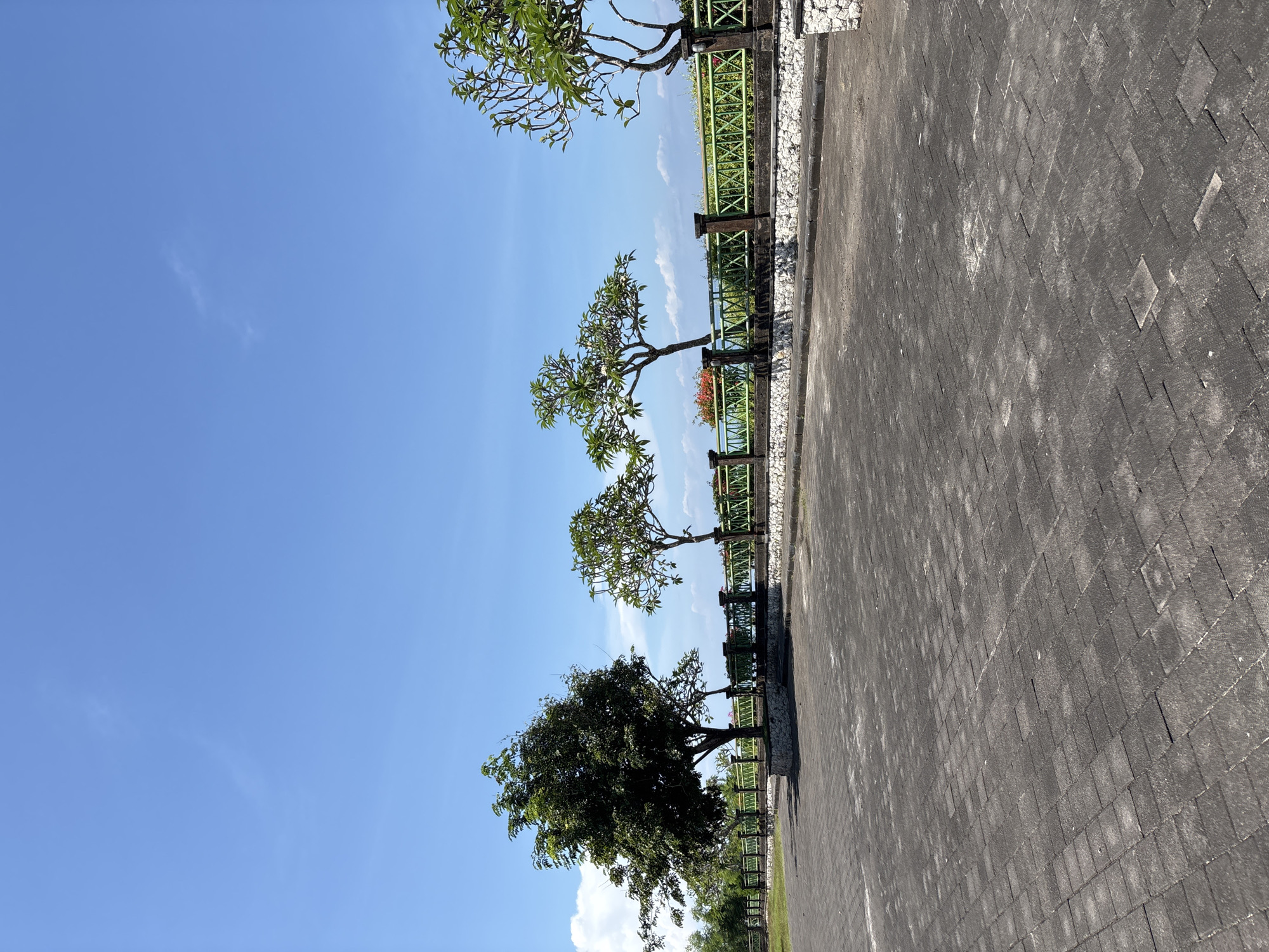 The image depicts a paved walkway in Kuta Selatan, Indonesia, bordered by a low green fence and a series of trees. The walkway is constructed of dark gray bricks, and the fence is a light green color. The trees vary in size and foliage density, with the smallest trees near the top of the image, and the largest near the bottom. The sky is bright blue with a few scattered clouds. Beyond the fence, a hint of the ocean can be seen.
