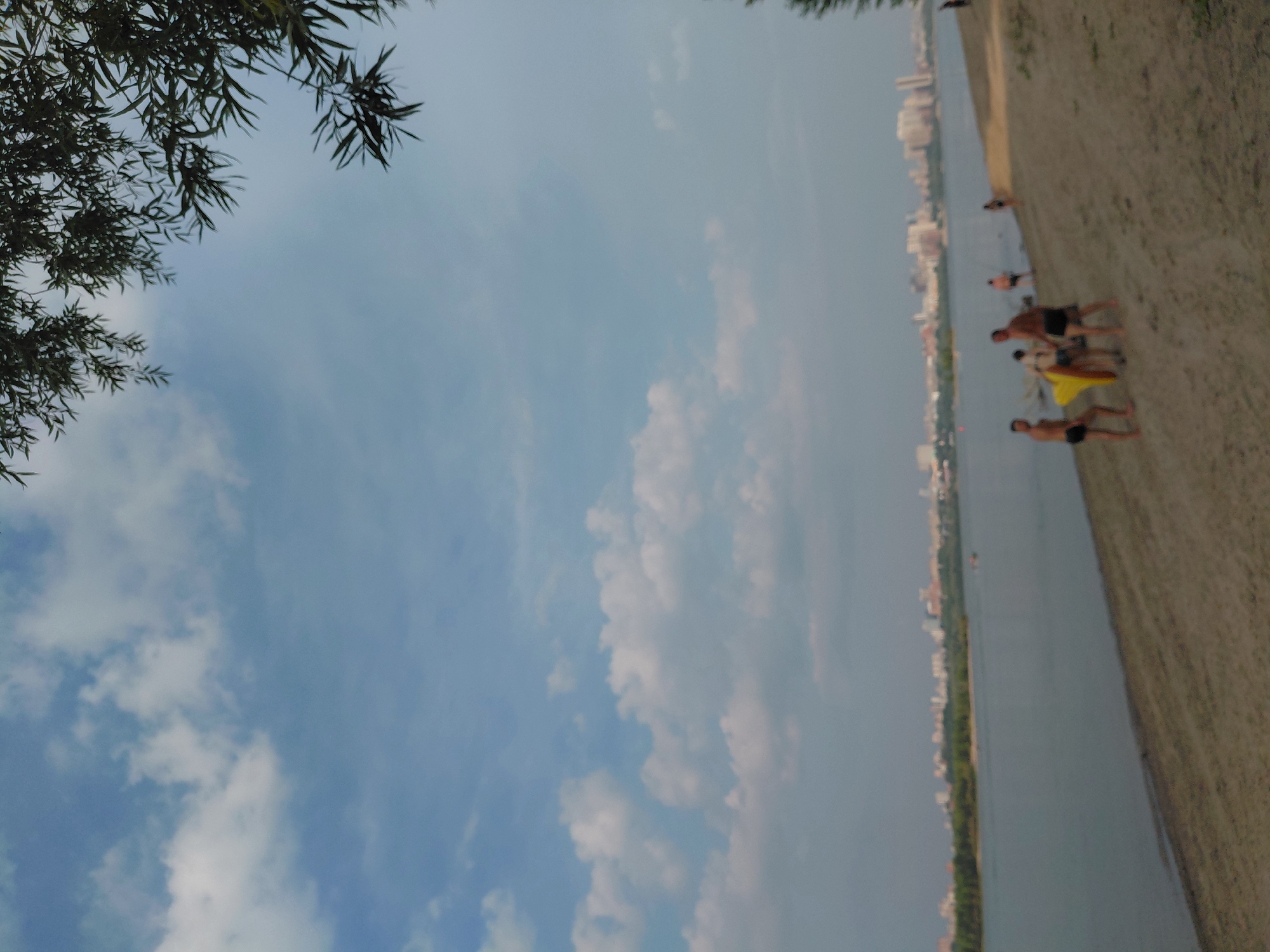 The image depicts a wide riverbank in a city, likely Novosibirsk, Russia, given the skyline. The sky is partly cloudy with a light blue hue. In the foreground, a sandy beach slopes down to the river. Several individuals are present on the beach. On the left side, three men are standing, one of whom is holding a yellow inflatable object. Further to the right, another man is walking, and a person, possibly a woman, is seen further down the beach near the water. In the background, across the river, a city skyline is visible, characterized by buildings of various heights and a lush green treeline separating the city from the water. The river itself appears calm, reflecting the sky. The overall atmosphere suggests a leisure activity on a warm day.