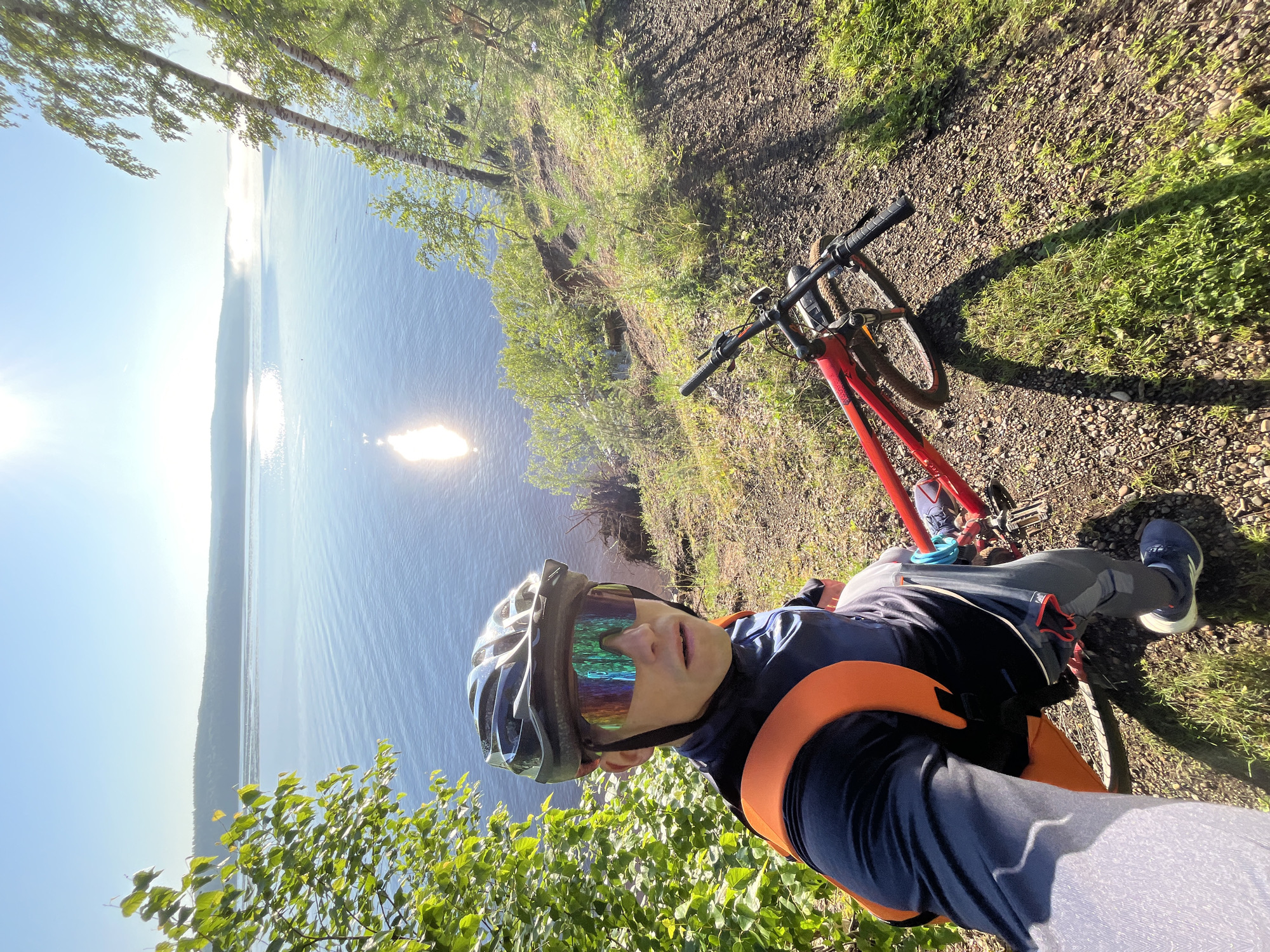 An outdoor, eye-level, selfie-style photograph shows a person wearing a helmet and colorful sunglasses, with a red bicycle parked beside them on a dirt path. The person is looking towards the camera with a neutral expression. Behind them, a calm body of water stretches towards a hazy horizon with a clear blue sky above, illuminated by the sun's bright reflection on the water. The immediate surroundings are hilly and covered with grass and trees. This location is the embankment of the Kama River in the city of Chaikovsky, Russia, known for its scenic views.