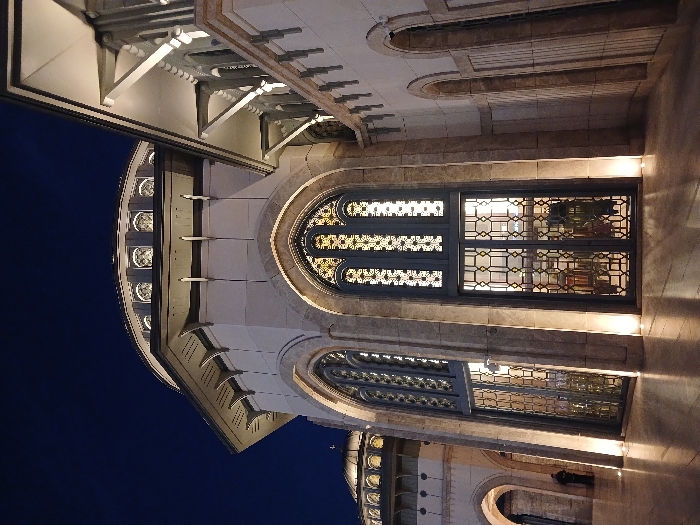 The image shows a portion of the exterior of the Camlica Mosque in Istanbul, Turkey. The mosque is a large, modern structure with a white facade and ornate detailing. The scene is captured at night and the mosque is illuminated, showcasing its intricate architectural details, particularly the large arched doorway with intricately patterned iron gates. The doors are closed and no people are visible. The image focuses on the mosque's facade, emphasizing its grandeur and architectural beauty.