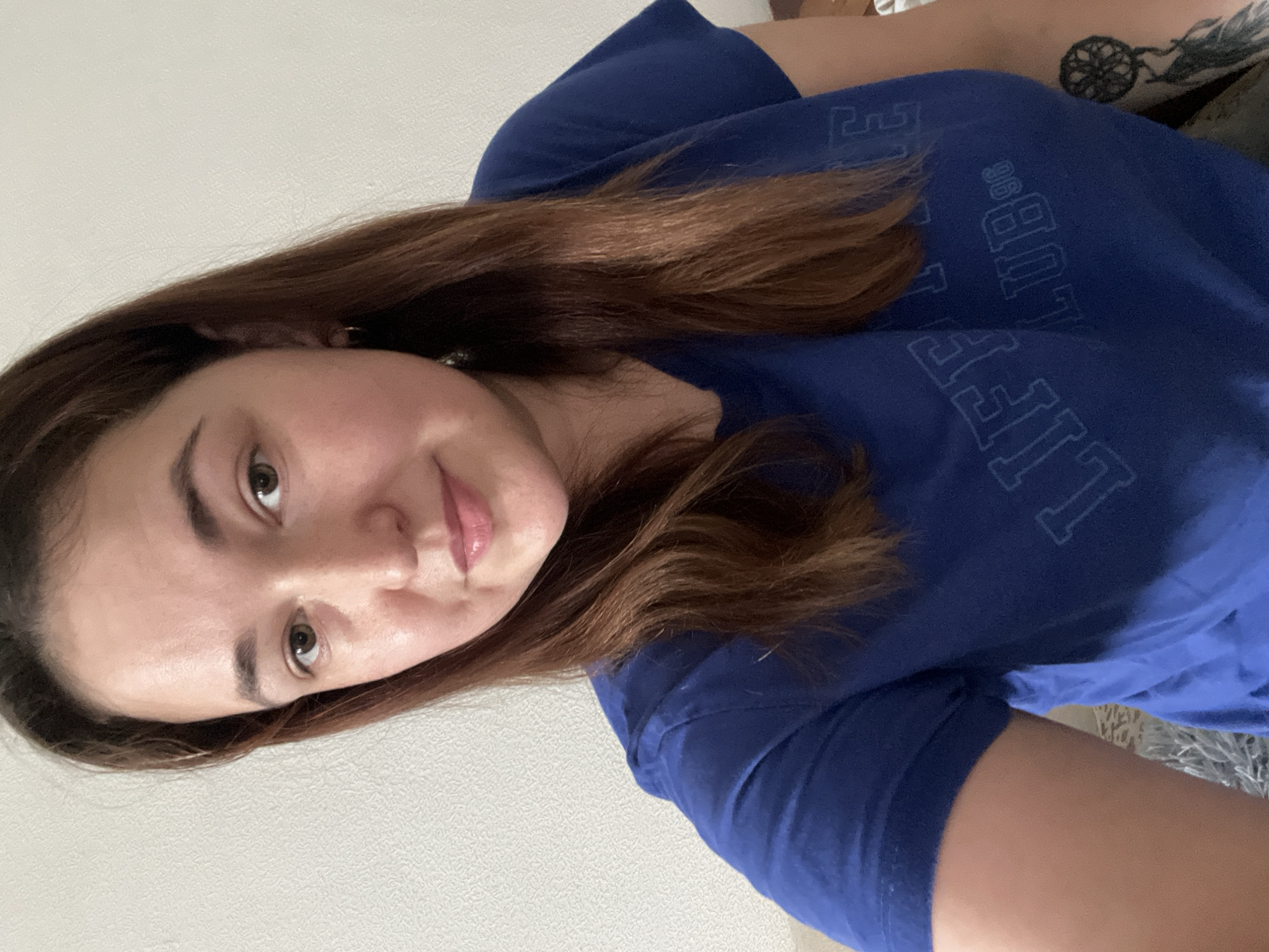 A woman is taking a selfie in front of a white textured wall. She has medium-length, reddish-brown hair and is wearing a blue t-shirt with the words "LIFE CLUB '96" printed on it. She has a dreamcatcher tattoo on her right arm. The image appears to be taken indoors, likely in a residential setting. There is no information available to indicate the location in Chelyabinsk, Russia.