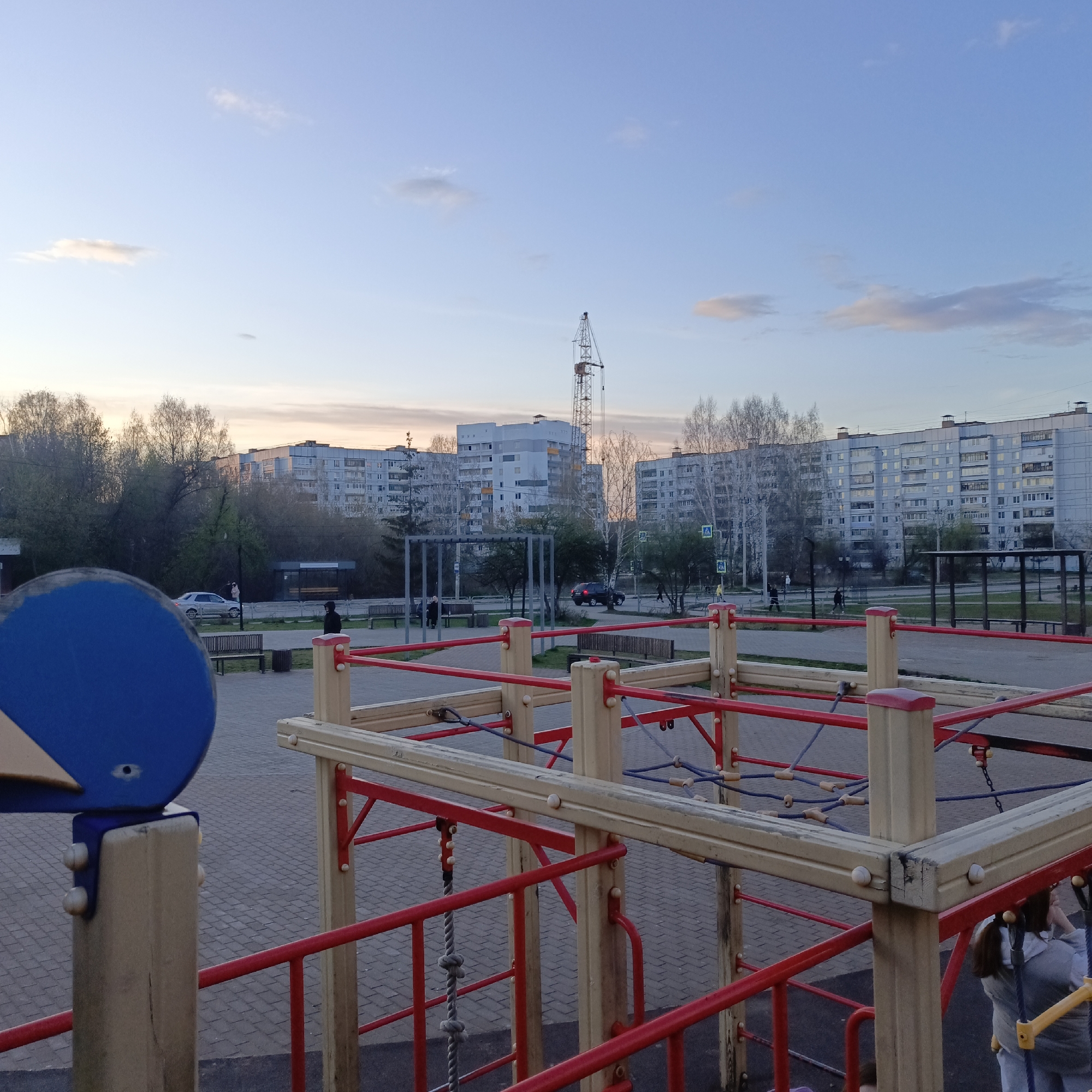 The image depicts an outdoor urban or suburban scene, primarily focusing on a children's playground in the foreground with residential buildings and a street in the background.
In the immediate foreground, a multi-level playground structure is visible, constructed from light-colored wooden beams and bright red metal bars. Blue ropes or nets form climbing elements within the structure, and part of a large blue circular or semi-circular playground element is prominent on the left. The ground in this area is paved with grey and brown interlocking bricks. A person, possibly a child, is partially visible at the bottom right, appearing to be on or near the playground.
The midground features a paved path or square, lined with benches and trees, many of which appear bare, suggesting early spring or late autumn. A silver car and a dark SUV are parked or driving on a road beyond the trees. A bus stop shelter with a clear roof and benches is also visible. Several people are scattered throughout the midground, walking along the paths or standing near the road. Blue and white pedestrian crossing signs (featuring a letter "A" symbol) are discernible near the street.
The background is dominated by several multi-story residential apartment buildings, predominantly light grey or white, with numerous windows. A tall construction crane stands prominently behind the buildings, indicating ongoing development in the area. The sky above is a soft gradient of light blue, transitioning to warmer, slightly orange and pink hues near the horizon, suggesting either dawn or dusk. There are a few wispy clouds scattered across the sky. The overall atmosphere appears calm and peaceful. No discernible text is visible on any signs or buildings beyond the symbols on the pedestrian crossings.
