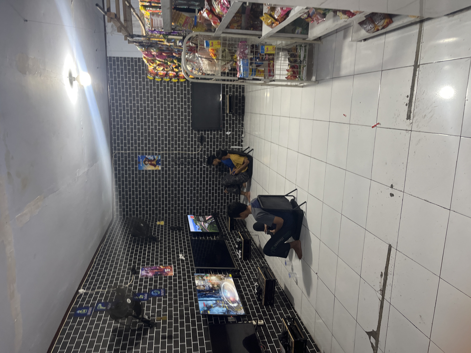 The image depicts an indoor gaming area, likely a small internet cafe or a dedicated gaming room, located in Laban, Indonesia. The room is characterized by dark, brick-patterned wallpaper and a white tiled floor. In the foreground, two young men are engaged in playing video games. One individual, wearing a gray and blue shirt and dark pants, is seated on a black gaming chair with a controller in his hands, facing a television screen displaying a racing game. Another individual, wearing a yellow shirt and patterned pants, is seated on the floor further back, also appearing to play a game.
Multiple television screens are mounted on the wall, with at least two actively displaying game content. Suspended from the ceiling are what appear to be fans. On the right side of the image, a metal shelving unit is stocked with various snack items, suggesting the establishment may also function as a small convenience store. The lighting in the room appears to be from overhead fixtures, one of which is prominently visible and illuminated. The overall environment suggests a casual and functional space designed for entertainment.