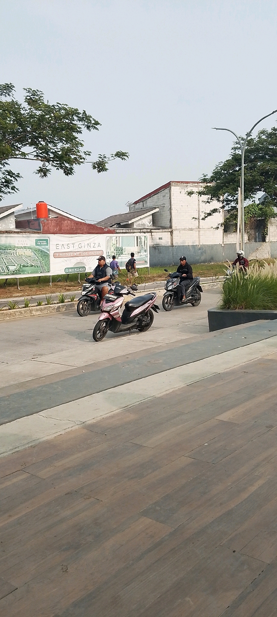 This is a medium shot, eye-level, outdoor, daytime photograph of a street scene in Satriamekar, Indonesia. The foreground is dominated by a wooden deck or walkway, with textured, plank-like surfaces. Beyond this, a paved road is visible, with several motorbikes and riders. One pink scooter is prominently displayed in the middle ground, with two other motorbikes and riders to its left and right. To the left of the road, a large banner advertising "EAST GINZA" is displayed, with images of a housing complex and contact information. Behind the banner, several unfinished or partially constructed white brick buildings with red-tiled roofs are visible. A large orange water tank is mounted on one of the buildings. To the right of the road, a street lamp with a curved arm stands tall, and lush green grass and bushes are visible, as well as a dark, raised planter box. In the background, trees with green foliage frame the top of the image, and the sky is a hazy, pale blue. There are several individuals on motorbikes and walking in the scene.