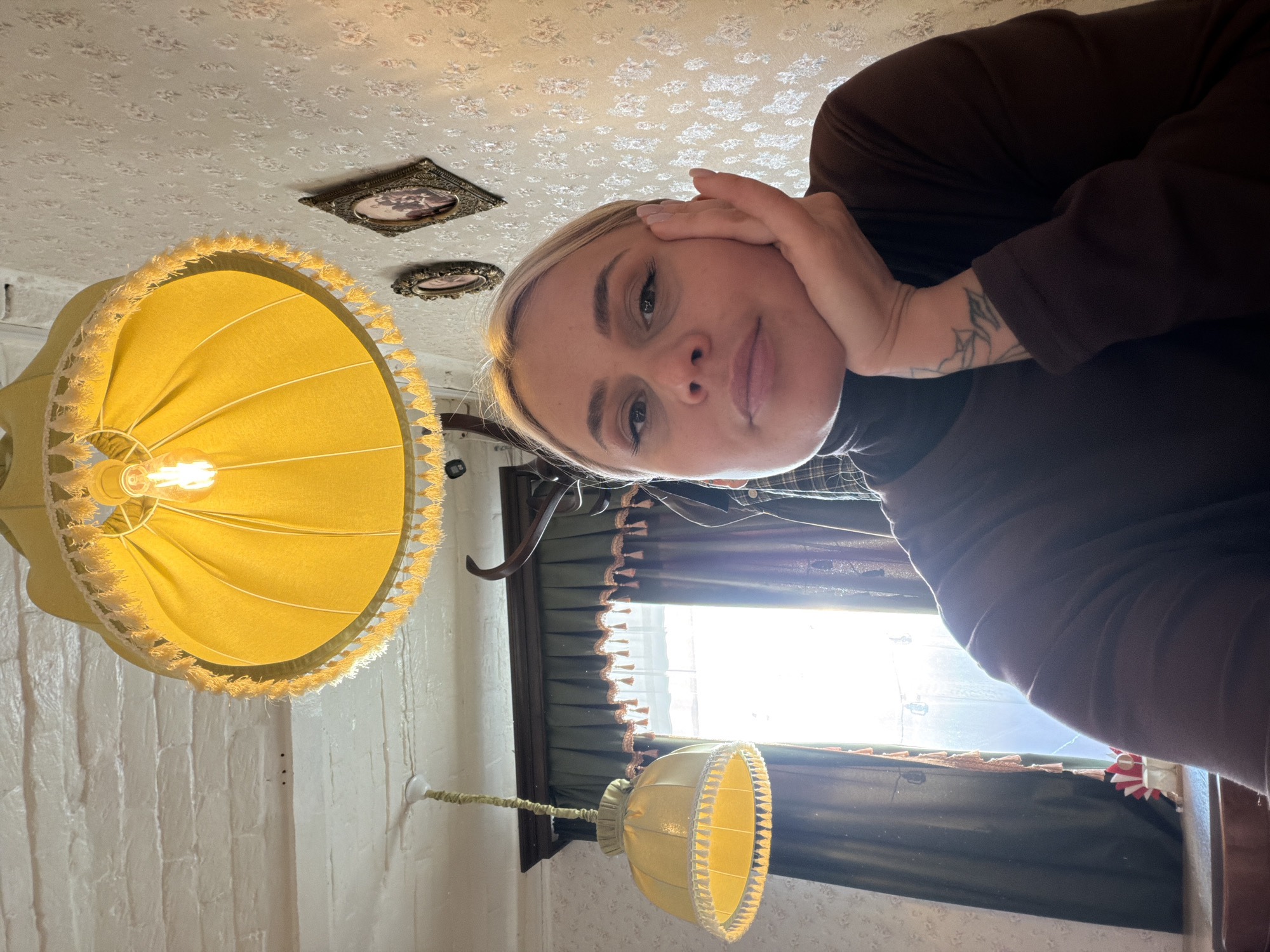 A woman with short blonde hair and a tattoo on her forearm is positioned in the foreground, holding her hand to her face. She is wearing a brown turtleneck shirt. The room appears to be indoors, with a low ceiling covered in a patterned wallpaper. Hanging from the ceiling is a yellow lampshade with fringed edges, illuminated by a visible light bulb. Behind her, a window with a dark frame and curtains allows sunlight to enter the room. Another smaller yellow lampshade is also visible near the window. Two small, framed pictures are affixed to the ceiling. Based on the information provided, the location is Sarapul, Russia.