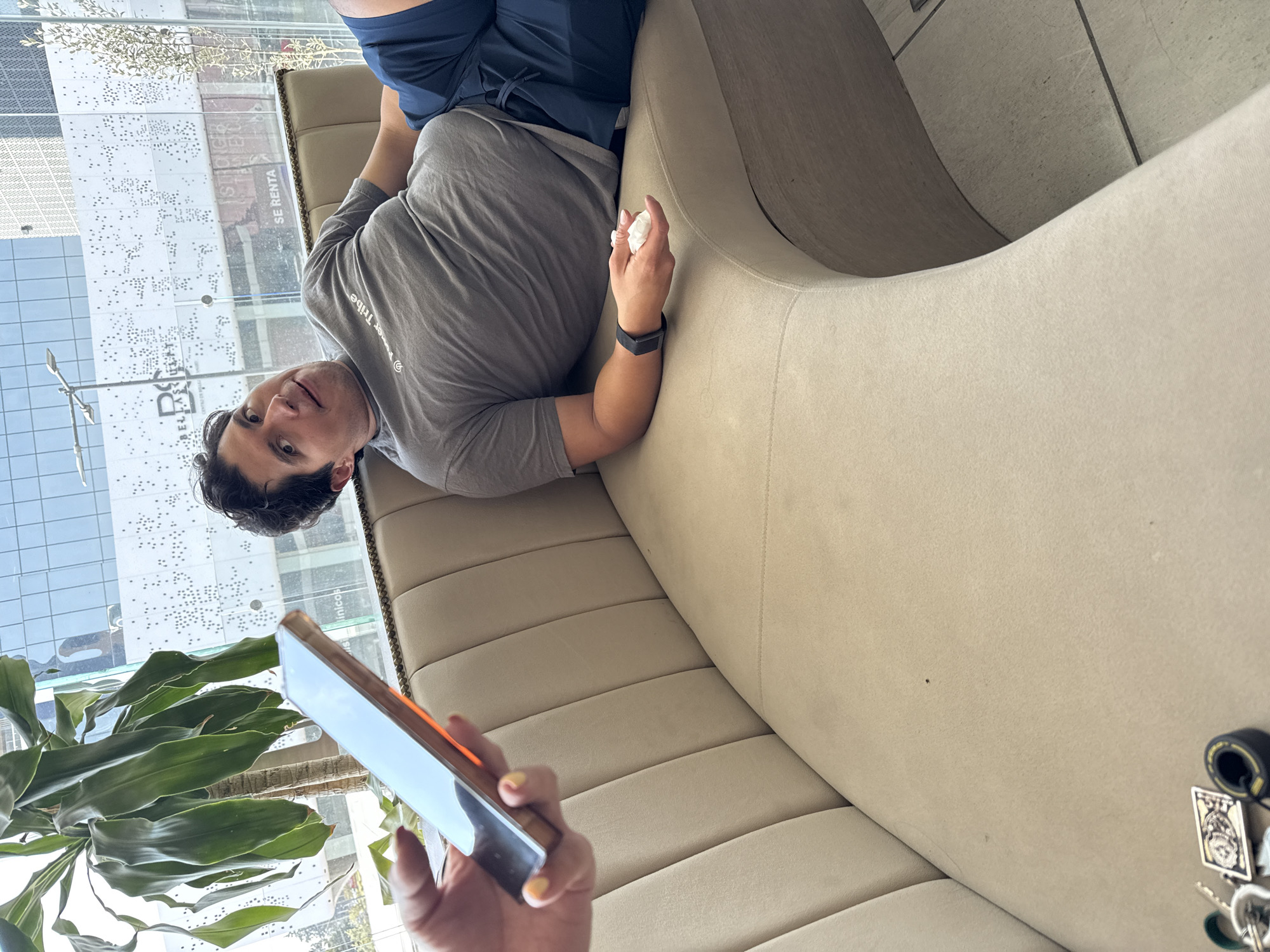 The image, correctly oriented, captures an indoor scene, likely a modern lounge or waiting area, looking out onto a city street in Puebla, Mexico.
A young man with dark hair and a neutral expression is reclining on a plush, beige, curved sofa. He is wearing a grey t-shirt, partially revealing the text "e Tribe" on his chest, and dark shorts or athletic wear. A black smartwatch is visible on his left wrist, and he holds a crumpled white tissue in his right hand. He looks directly towards the camera.
In the immediate foreground, a person's hand, with yellow-painted fingernails, holds a smartphone with a gold/orange metallic frame and a dark screen, appearing to take the photograph.
Behind the man, expansive windows reveal a bright, sunny daytime environment. Outside, modern buildings are visible; one features a distinctive facade with numerous small dark dots, while another has partially visible text "SE RENTA" (Spanish for "FOR RENT") and "DEL LAS". A large green leafy plant is situated near the window on the left. On the right side of the sofa, a set of keys attached to a distinctive rectangular keychain, which features a detailed illustration of a face or skull, is resting. The overall mood is casual and relaxed.