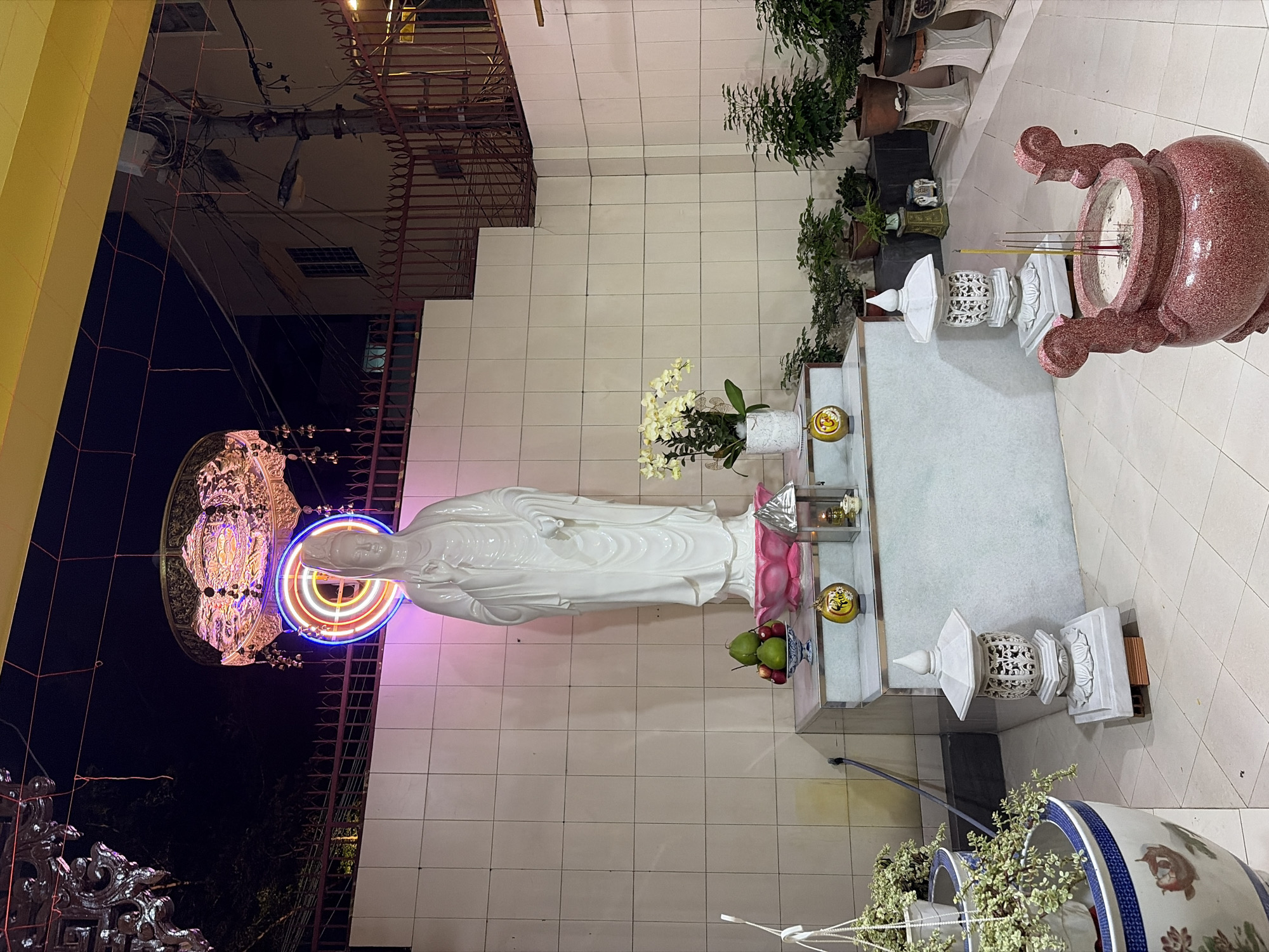 This image depicts an outdoor shrine at night in the city of Thủ Đức, Vietnam. The focal point is a tall, white statue of a female deity, likely Quan Âm (Goddess of Mercy), standing on a tiered pedestal. The statue is adorned with a pink lotus base. In front of the statue, on the pedestal, is a bowl of green and red fruits, a white potted orchid, and a small golden orb with Chinese characters. To the left and right of the pedestal are decorative white lanterns. In the foreground, a large, ornate red incense burner with a speckled finish contains burning incense sticks. Various potted plants, including a large one with trailing vines to the left, are arranged around the shrine. Behind the statue, a tiled wall rises, topped by a metal fence. Above the statue, a circular neon light fixture with blue and pink hues and a more ornate metallic light fixture hang. The background shows a dark sky with some electrical wiring and buildings visible. The ground is tiled.