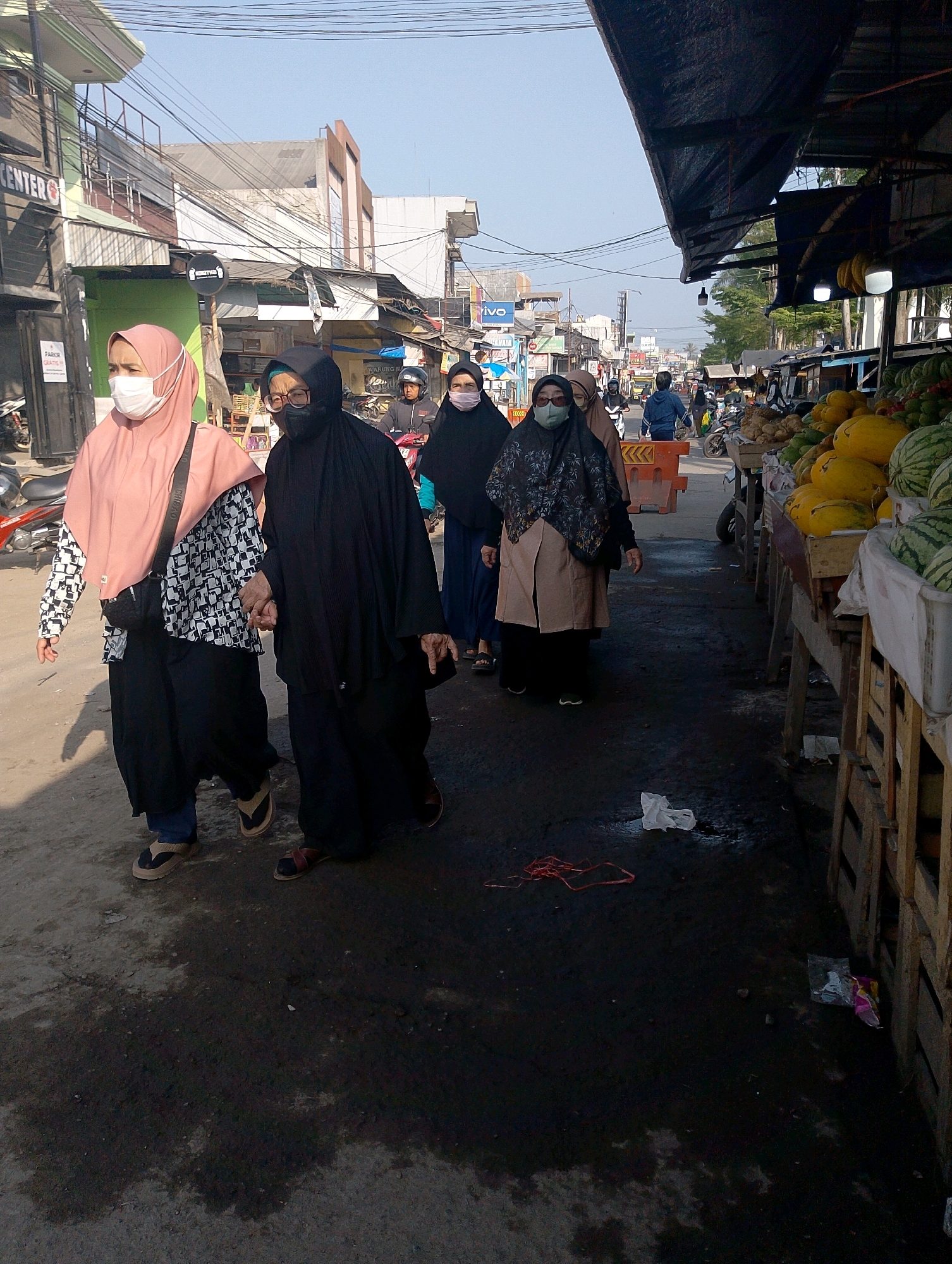 The image depicts a street scene in Lembang, Indonesia, likely a marketplace, under a clear sky. The foreground shows several individuals, mostly women, walking on a paved surface that appears wet in places. Two women in the foreground are holding hands; one wears a light pink hijab and a patterned top with black pants, while the other is fully covered in black attire. They are both wearing masks. Behind them, other individuals are also walking, some wearing hijabs and masks.
To the right, a market stall displays a variety of fruits, including watermelons and what appear to be melons or gourds. The stall is constructed of wood and covered with a dark tarp. In the background, there are buildings lining the street, some appearing to be shops or businesses with signage. Power lines are visible crisscrossing overhead. A few motorcycles and a person in a blue jacket are also present in the middle ground, suggesting a moderately busy street. The overall environment is consistent with a daytime scene in a tropical or subtropical urban area.