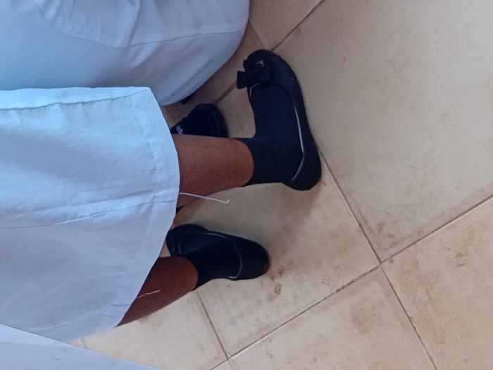 The scene in Ejule, Nigeria, displays the lower extremities of at least two individuals on a tiled floor. One person, positioned on the left, shows bare lower legs with a dark skin tone, wearing black ankle socks and plain dark flat shoes. A light-colored garment, possibly a skirt or trousers, covers the upper portion of their legs, with a loose white thread hanging from its hem. To the right, part of a second individual is visible, also wearing a light-colored garment, black ankle socks, and dark flat shoes, with the right shoe featuring a small decorative bow. The floor is composed of light-beige square tiles with dark grout lines, exhibiting minor smudges and discoloration.