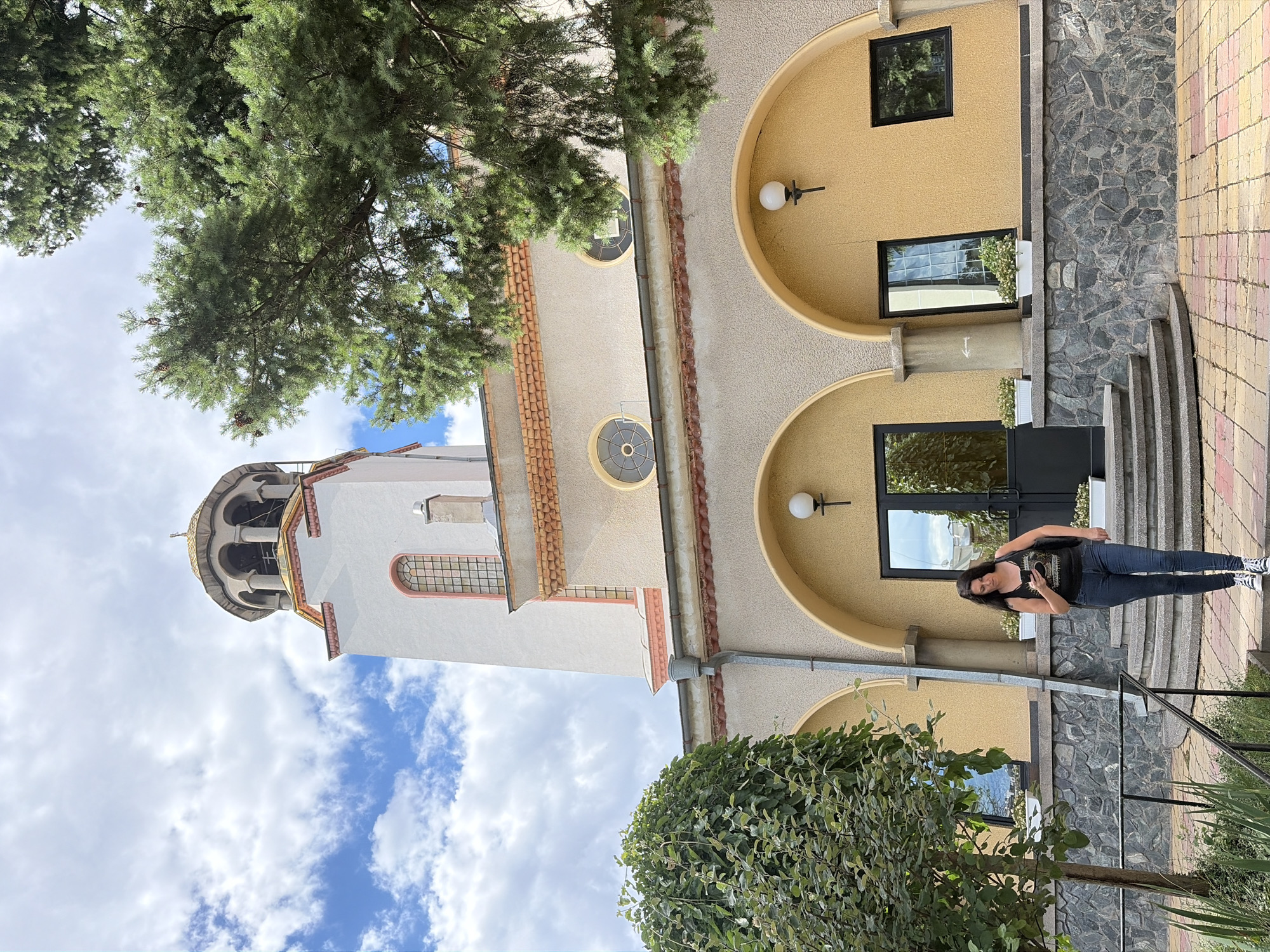 A woman is walking down a set of concrete stairs in front of a church in Sofia, Bulgaria. She is wearing a black tank top and blue jeans, and is looking at her phone. The church has a tall white bell tower with a golden dome, and a yellow facade with arched windows and doorways. There are trees and bushes around the church, and the sky is blue with white clouds. The ground is paved with colorful tiles.