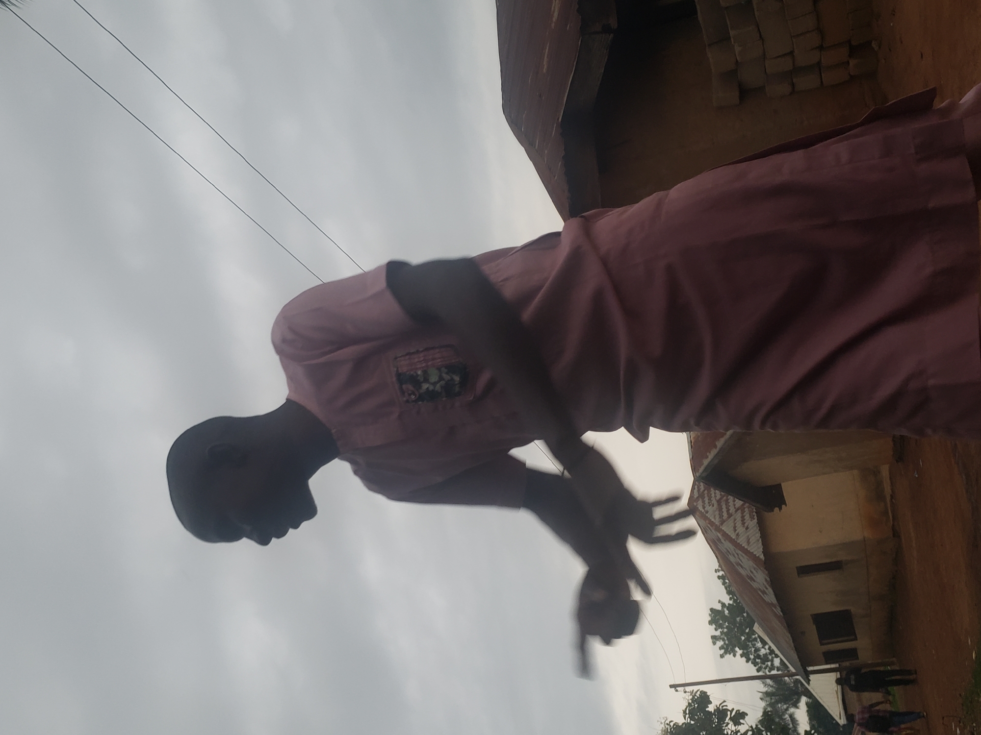 In Gumel, Nigeria, under a cloudy sky, a person stands outdoors, captured in silhouette against the overcast atmosphere. They are wearing a pink, ankle-length traditional garment with short sleeves and a detailed pattern on the chest pocket. Their head is turned to the left, with facial features obscured by shadow. Their right arm is extended forward, with their hand pointing, while their left arm is bent at the elbow and also extended. Behind the person, two simple, single-story structures with corrugated metal roofs are visible. The ground is reddish-brown earth. In the distance, another building and trees can be seen, along with a utility pole. The image appears to be taken from a low angle, emphasizing the figure against the sky.
