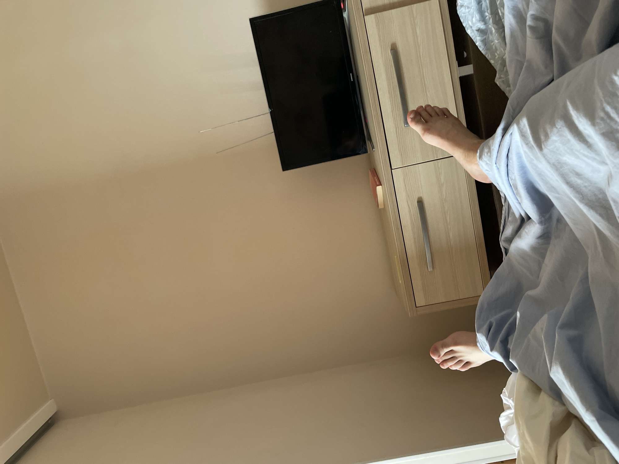 The image depicts a bedroom scene. In the foreground, a person's feet are visible, emerging from under light blue bedding. The feet appear to be resting, suggesting the person is in bed. To the right of the bed, there is a wooden dresser with drawers, and on top of the dresser, a flat-screen television is placed, with an antenna extending upwards. To the left of the television, a small book is visible on the dresser. The walls of the room are painted a neutral, light beige color. The scene is captured from a slightly elevated perspective, looking down towards the bed and dresser. There are no distinct architectural features or cityscapes visible that would indicate a specific location in Kyiv, Ukraine.
