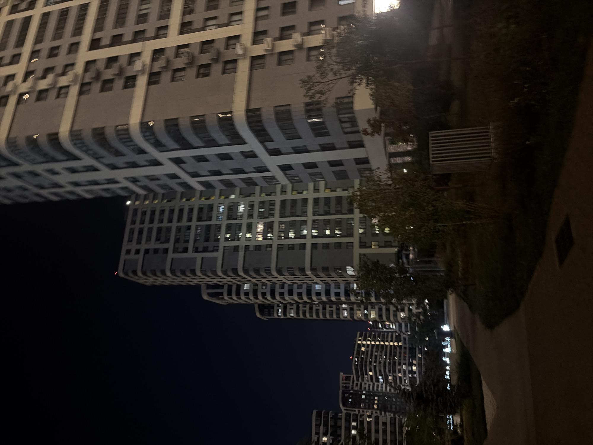 This image captures a nighttime view of a modern residential complex in Moscow, Russia. Tall, multi-story apartment buildings dominate the skyline, their facades illuminated by numerous interior lights, indicating occupancy. The architecture is characterized by curved balconies and a predominantly grey and white color palette. In the foreground, a paved pathway winds through a dimly lit park area. Sparse trees and vegetation are visible, with some streetlights casting a soft glow. A utilitarian, slatted metal structure, possibly a trash receptacle or ventilation unit, is situated in the park. The sky is dark, characteristic of nighttime in an urban environment.
