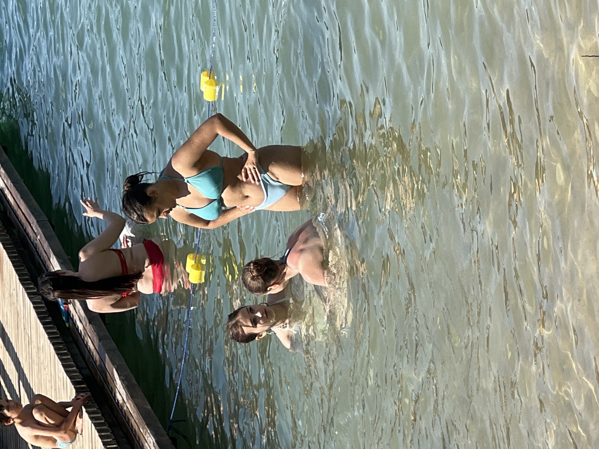 In Vilnius, Lithuania, a group of young people are enjoying a sunny day at a lake. The image captures a candid moment as they stand in the clear, shimmering water near a wooden dock. The water is shallow enough for them to stand comfortably, and the sunlight creates a dappled effect on the surface.
Several individuals are visible in the water, all wearing swimwear. In the foreground, a person in a red bikini is partially submerged, with their long dark hair cascading around them. To their left and slightly behind, another person in a light blue bikini is looking towards the viewer with a smile. Further into the water, two more individuals are visible, one in the background with their back to the camera and another in the mid-ground facing away from the camera. A young boy is seated on the wooden dock in the lower left corner of the frame, appearing relaxed.
The scene suggests a leisurely summer day, with the bright sunlight and clear water indicating fair weather. The wooden dock suggests a recreational area, possibly a beach or a designated swimming spot. The overall mood is one of carefree enjoyment and relaxation. There is no visible text in the image.