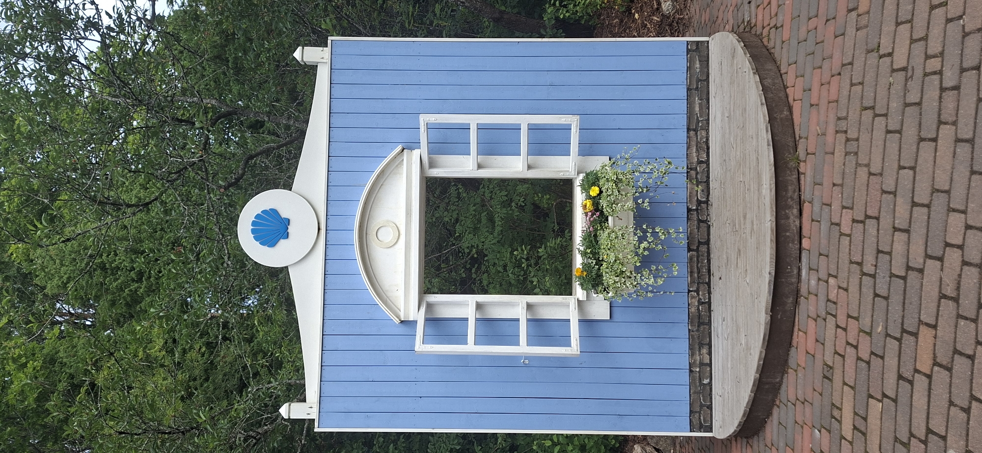 The image shows a brightly colored, artificial facade with a blue wooden wall, white trim, and a decorative blue shell emblem at the top. The facade features a white-framed window with an open top sash, and a flower box beneath it containing green trailing plants and yellow and pink flowers. The facade is situated on a wooden stage with a curved edge, which is surrounded by a brick pathway. Behind the facade, a dense green forest is visible through the window. The scene is outdoors, suggesting a park or public space. This facade is located in the city of Naantali, Finland, specifically at the entrance to the Moomin World theme park.