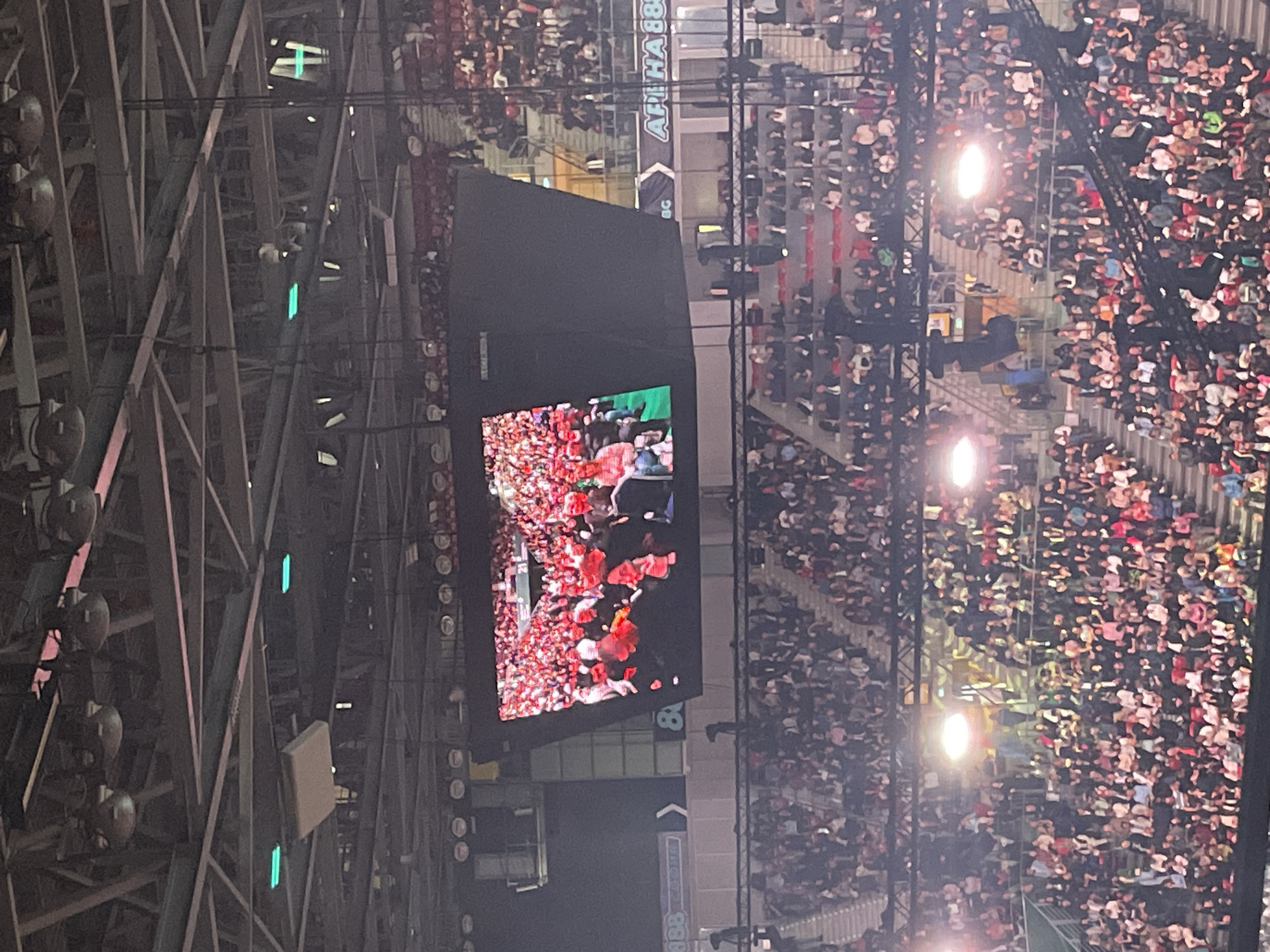 This image captures a large indoor arena or stadium, densely populated with a vast crowd of spectators, suggesting a major event is underway. The perspective is from an elevated position, looking down across one section of the venue.
A prominent feature is a large video screen, centrally positioned, displaying dynamic content. On the screen, a vibrant scene unfolds, showing what appears to be a performer or group amidst a flurry of red elements, possibly confetti, petals, or light effects, with some green elements in the background.
The tiered seating areas are packed with people, illuminated by various overhead lights and spotlights. The arena's infrastructure includes extensive metal truss systems that support the roof, various lighting fixtures, and what appear to be sound system components. Some green strip lights are also visible within the metal framework.
The setting is clearly nighttime or evening due to the reliance on artificial lighting throughout the venue. A banner is visible in the upper right section of the image, displaying the text "APHA 88BG," which provides a potential location cue for Sofia, Bulgaria. The overall atmosphere suggests an energetic concert, sporting event, or similar large-scale performance.