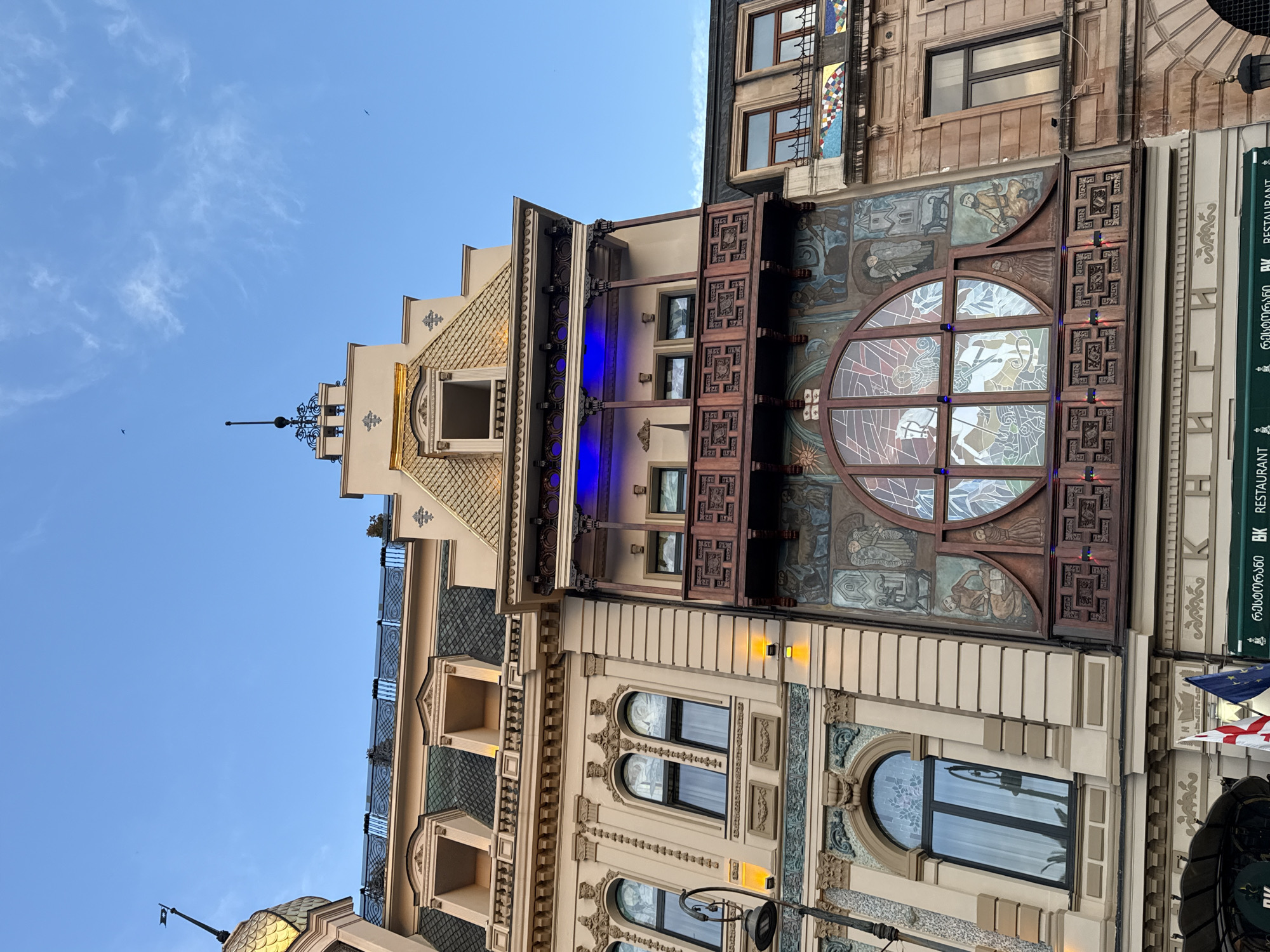 The image displays a detailed upward view of the ornate facades of three adjacent buildings in Batumi, Georgia, against a clear blue sky with minimal wispy clouds.
The leftmost building features a light beige facade with classical architectural elements, including arched ground-level windows and rectangular windows with decorative surrounds on the floor above. Its roof is dark-tiled with dormer windows. A golden-scaled dome or cupola with a dark finial and a small dark pennant is visible on the far left.
The central building, also light-colored, is highly decorated. Its upper section has a steep, gold-tiled peaked roof topped with a dark, spired finial. Below this is an open dormer-style window. A dark wooden balcony adorned with intricate carvings and supported by columns projects outward; it is illuminated by blue lights from above. Below this, a larger, elaborately carved wooden facade features a prominent circular window divided into sections, possibly stained or painted glass, depicting abstract or illustrative designs. This window is flanked by painted murals on the building's surface, showcasing figures, animals, and architectural motifs in a traditional style. Small red and blue lights are integrated into the lower wooden carving of this facade.
The rightmost visible building has a simpler, brown-toned facade with rectangular windows and a band of colorful mosaic tiles near its top.
A black, ornate streetlight pole is partially visible on the far left. On the ground level, a green awning displays Georgian script "ხაჭაპურის სახლი" (Khachapuri's House) and "BK RESTAURANT". Partially visible flags, including the Georgian flag, are present below the central building's lower window. No individuals are visible. Two small, dark bird silhouettes are discernible in the clear sky.
