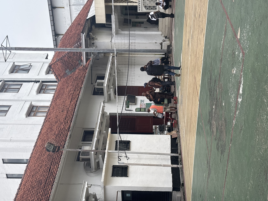 A group gathers on a green and beige painted court next to a white building with a red tile roof. Some individuals are seated, and others stand. A volleyball net stretches across the center. The building has multiple windows and air conditioning units. In the background, more people stand near an archway, and the sky is overcast.