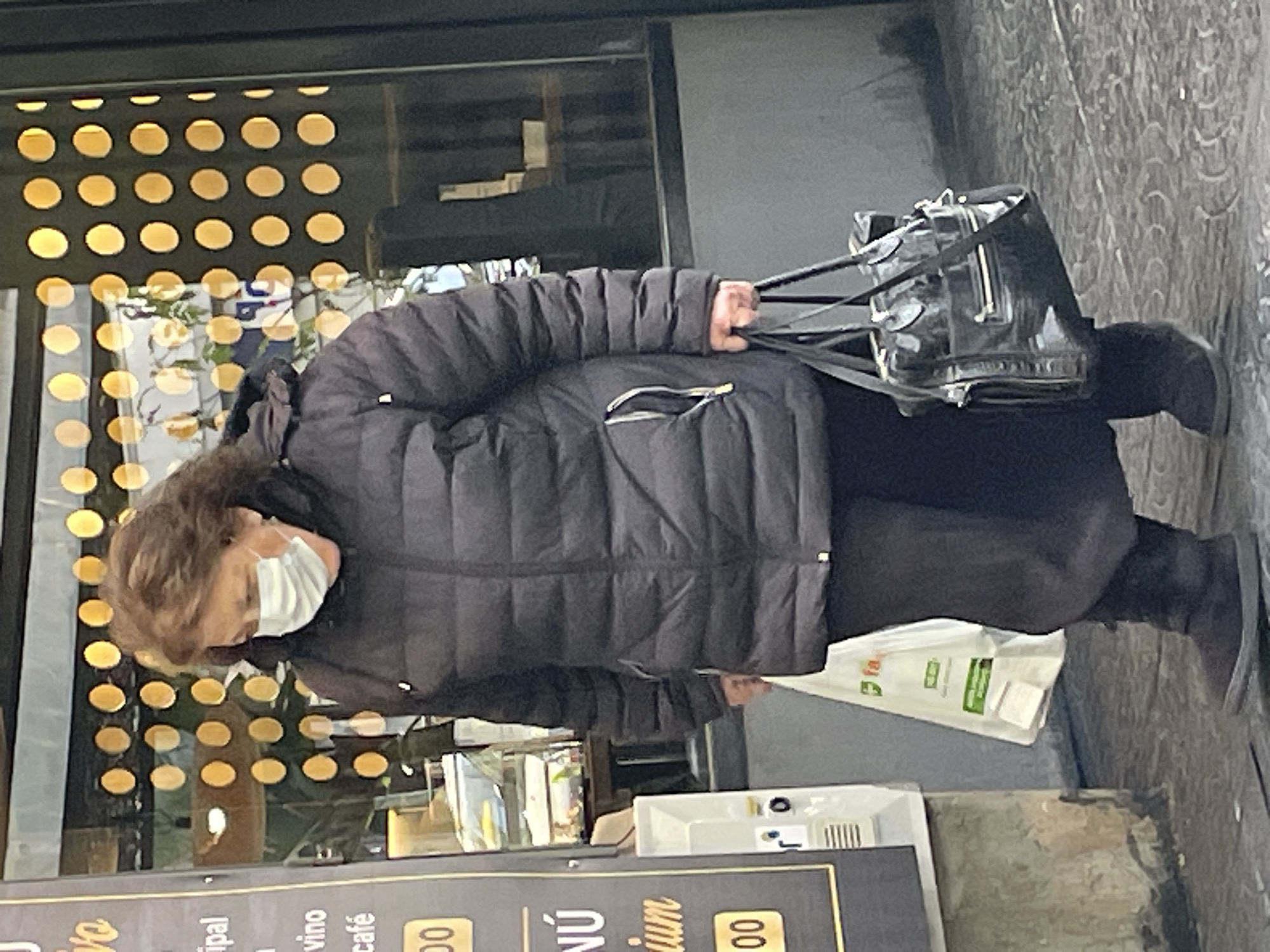 An elderly woman wearing a face mask, black padded jacket, and black pants walks on a paved sidewalk. She is holding a black leather purse and a white paper bag. The sidewalk borders a building with a menu board visible near the bottom of the frame. Through a glass window or facade with a grid of circular openings, a building interior is visible, likely a restaurant or cafe. The location appears to be an urban area in Buenos Aires, Argentina.