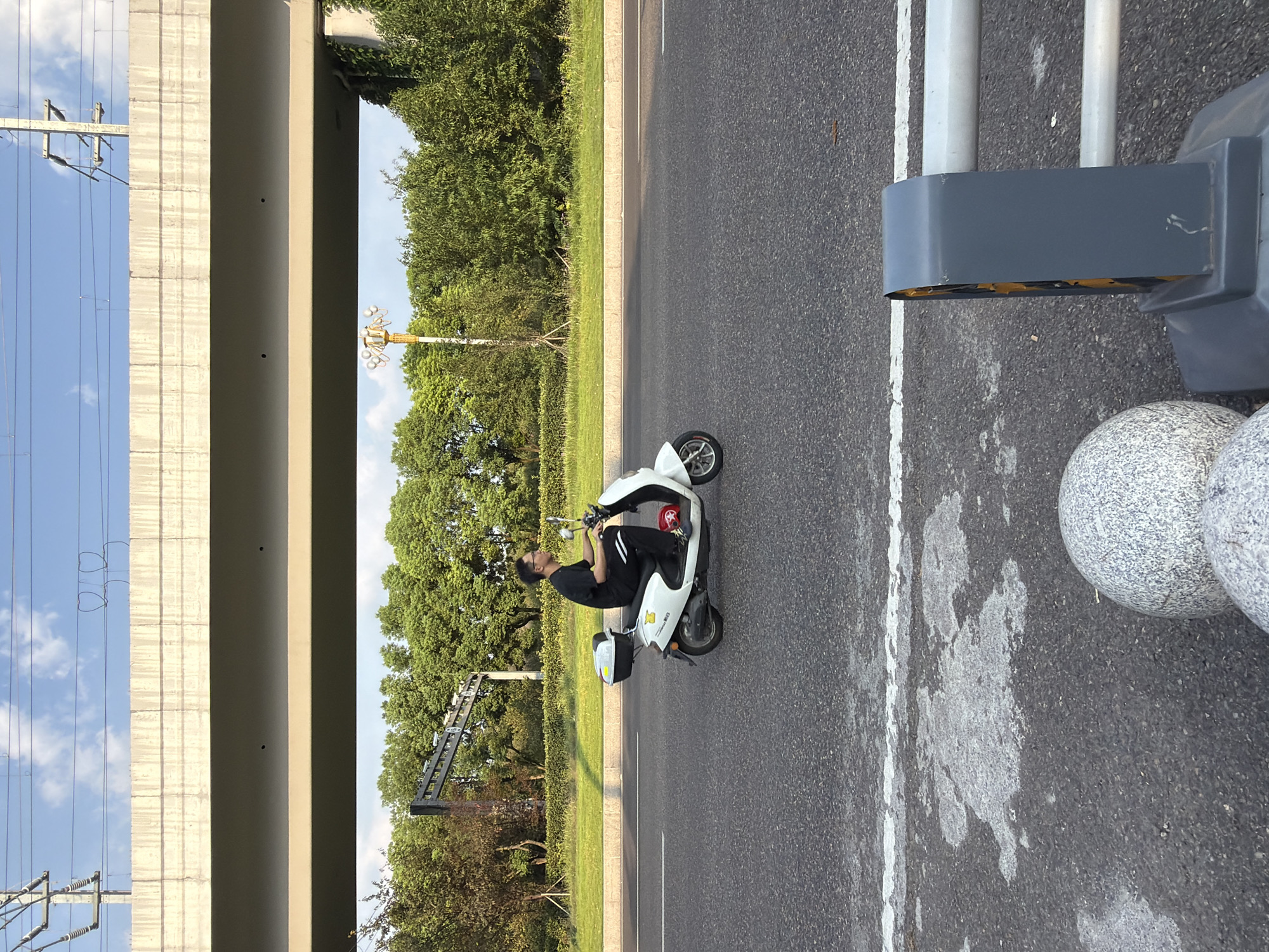 A man is riding a white electric scooter on a paved road in Jiangjin, China. The man is wearing a black t-shirt and black pants. He has glasses and short black hair. He is looking forward as he rides. The scooter has a black storage box on the back. A red helmet is on the floorboard of the scooter. In the background, there is a grassy area with trees and a highway overpass. The sky is blue with some white clouds. A traffic signal is visible in the foreground on the right side of the image. There are also two round, speckled bollards in the foreground.