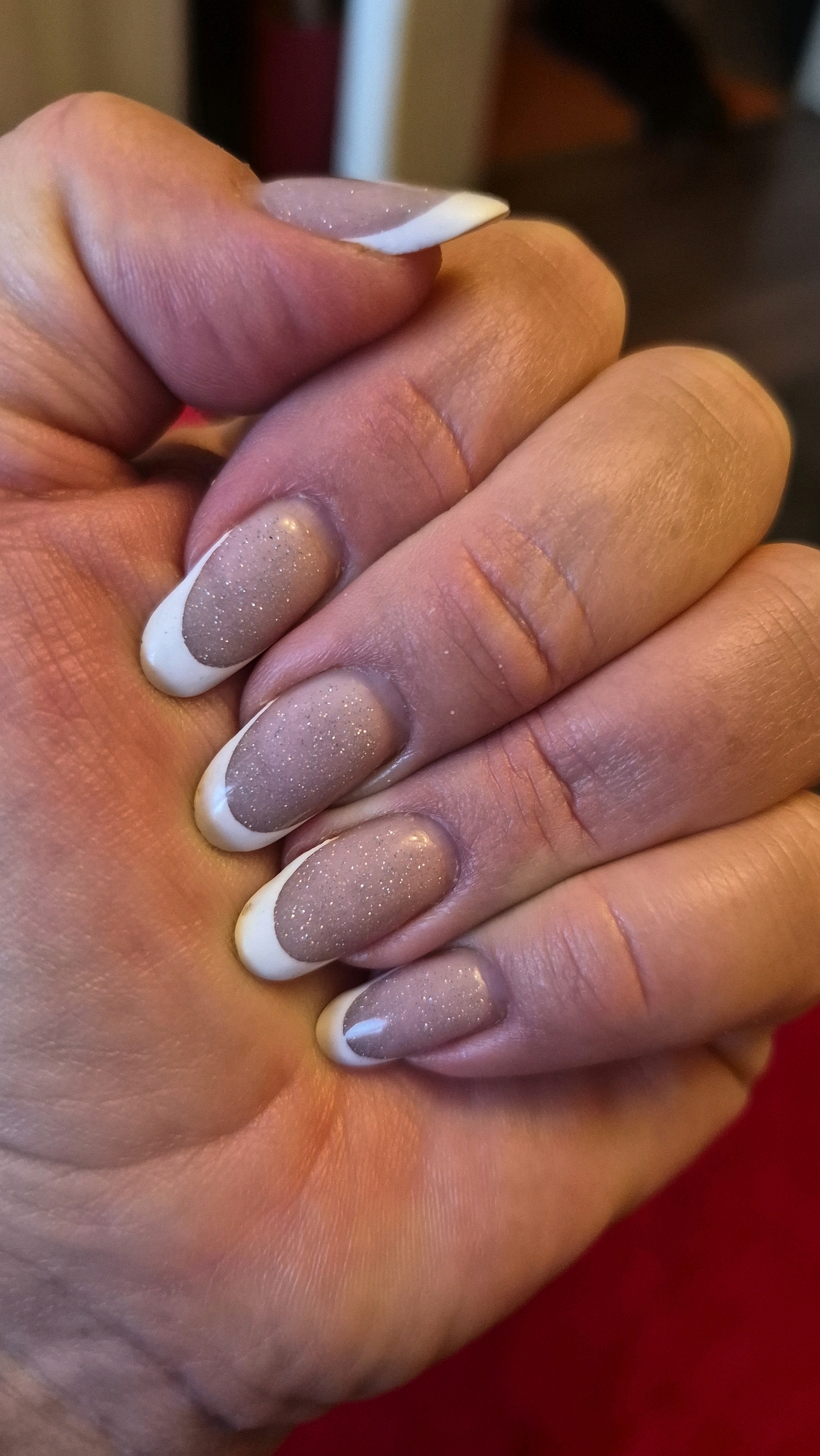 The image shows a close-up of a person's left hand with a french manicure. The fingernails are medium length and rounded, with a white tip and a subtle glittery nude base. The hand is resting on a red surface, and the background is blurred, suggesting an indoor setting. There is no indication of the location being in Krauja, Latvia. The image focuses solely on the hand and nails.