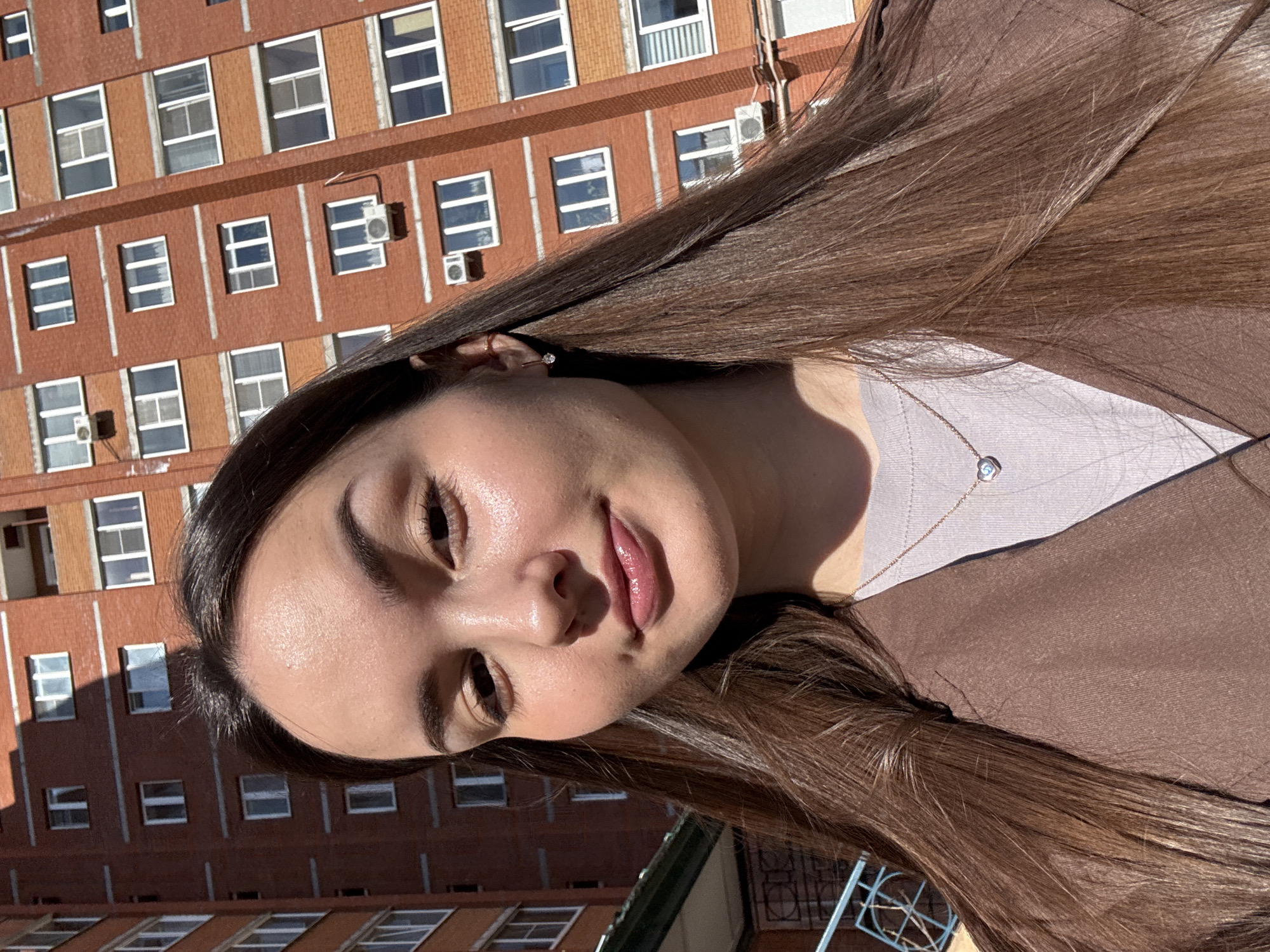 A young woman with long, brown hair is positioned in front of a large, brick building with numerous windows. She is wearing a light-colored top underneath a brown jacket. A delicate necklace with a small pendant is visible around her neck. The scene appears to be outdoors during daylight hours. The location may be inferred to be Chaikovsky, Russia.