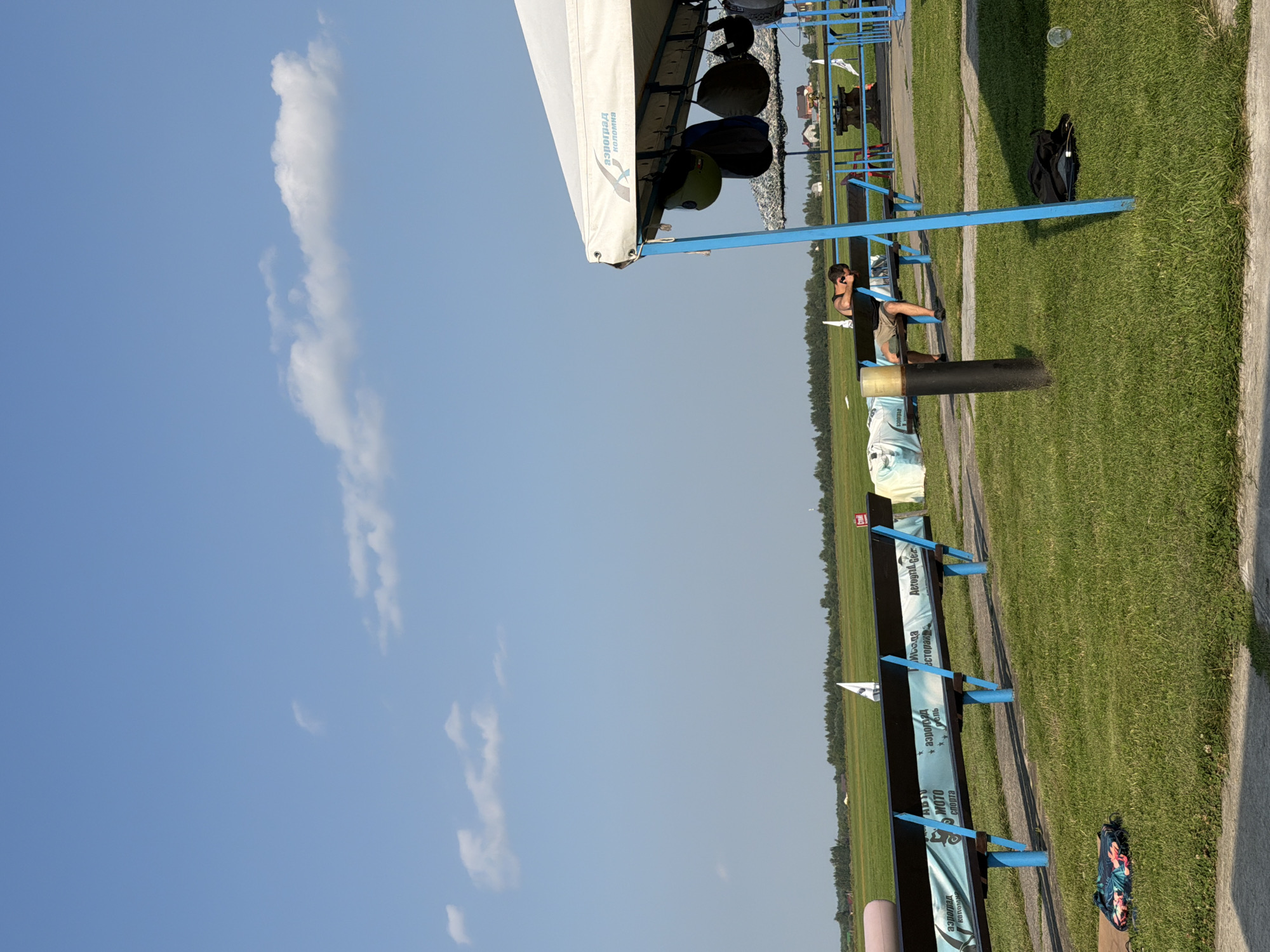 An outdoor scene shows a man in shorts and a dark tank top leaning his head on his arms on a blue-legged bench. The bench has a dark plank seat and backrest, with a banner advertising "Aerograd Kolomna" and "Avto Moto Sport" attached to its front. The bench is situated on a grassy area with a paved path in the foreground.
In the background, there's a wide expanse of green fields under a clear blue sky with scattered white clouds. Beyond the field, a line of trees forms the horizon. A white tent structure with "Aerograd Kolomna" printed on its fabric is visible on the right, with several helmets hanging from its supports. A black backpack lies on the grass near the tent. The location is identified as Korobcheevo, Russia.