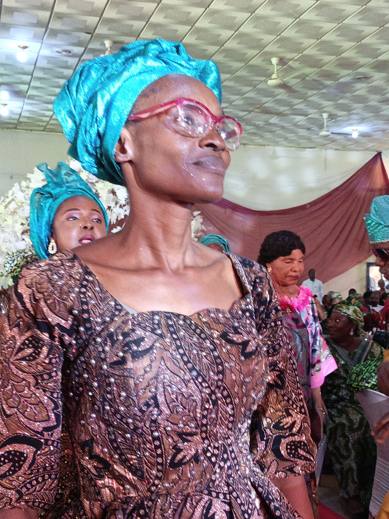 In an indoor setting, possibly in Fagge D2, Nigeria, the image captures several women dressed in vibrant, patterned clothing and headwraps. The main subject is a woman wearing a brown and black patterned dress adorned with small reflective embellishments. She has a bright blue headwrap and wears pink-framed glasses. Another woman in a matching headwrap is visible behind her, partially obscured, wearing earrings. In the background, other women are seen, including one with a floral necklace over a grey and pink patterned dress. The environment includes a tiled ceiling with ceiling fans and what appears to be a draped maroon curtain or backdrop. The overall impression is of a gathering or celebration.