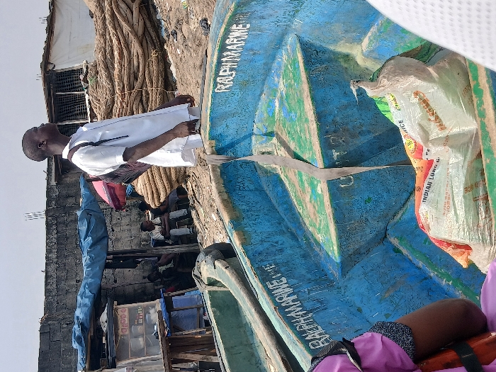 This image depicts a bustling waterfront scene in Alaiyabiagba, Nigeria, characterized by informal market activity and maritime elements under bright, hazy daylight.
In the foreground, the interior of a weathered, blue and green painted boat, likely wooden or fiberglass, occupies the lower central and right portions of the frame. The words "RALPH MARINE" are stenciled in white capital letters on its interior side. Several white sacks, one prominently displaying "INDIAN LONG GRAIN RICE," are placed as cargo within this boat. A grey strap is visible extending vertically from the boat's side down to its floor. Partially visible on the far left, a person wearing a purple garment and an orange life vest strap is seated in an adjacent, obscured vessel.
In the midground, a dark-skinned male, appearing to be a young adult, stands facing towards the upper right of the frame, his gaze directed upwards and away from the camera. He wears a short-sleeved white collared shirt, dark trousers, and a dark backpack on his left shoulder. He appears to be standing in or near a boat, or on the immediate shoreline. Behind him, on the cluttered ground, a makeshift market stall with a blue tarp roof is visible. The stall displays small packaged goods on shelves. Several individuals are seated in this area, some under the blue tarp, others further right under what appears to be a red umbrella or tarp. A substantial pile of thick, coiled ropes, possibly for fishing or mooring, is stacked on the ground to the right of the market stall.
The background consists of a grey concrete block wall, indicative of a permanent structure or boundary. Above and behind this wall, a few rudimentary structures with corrugated metal roofing are visible under a bright, overcast, or hazy sky. The immediate ground appears uneven, sandy, and scattered with debris.