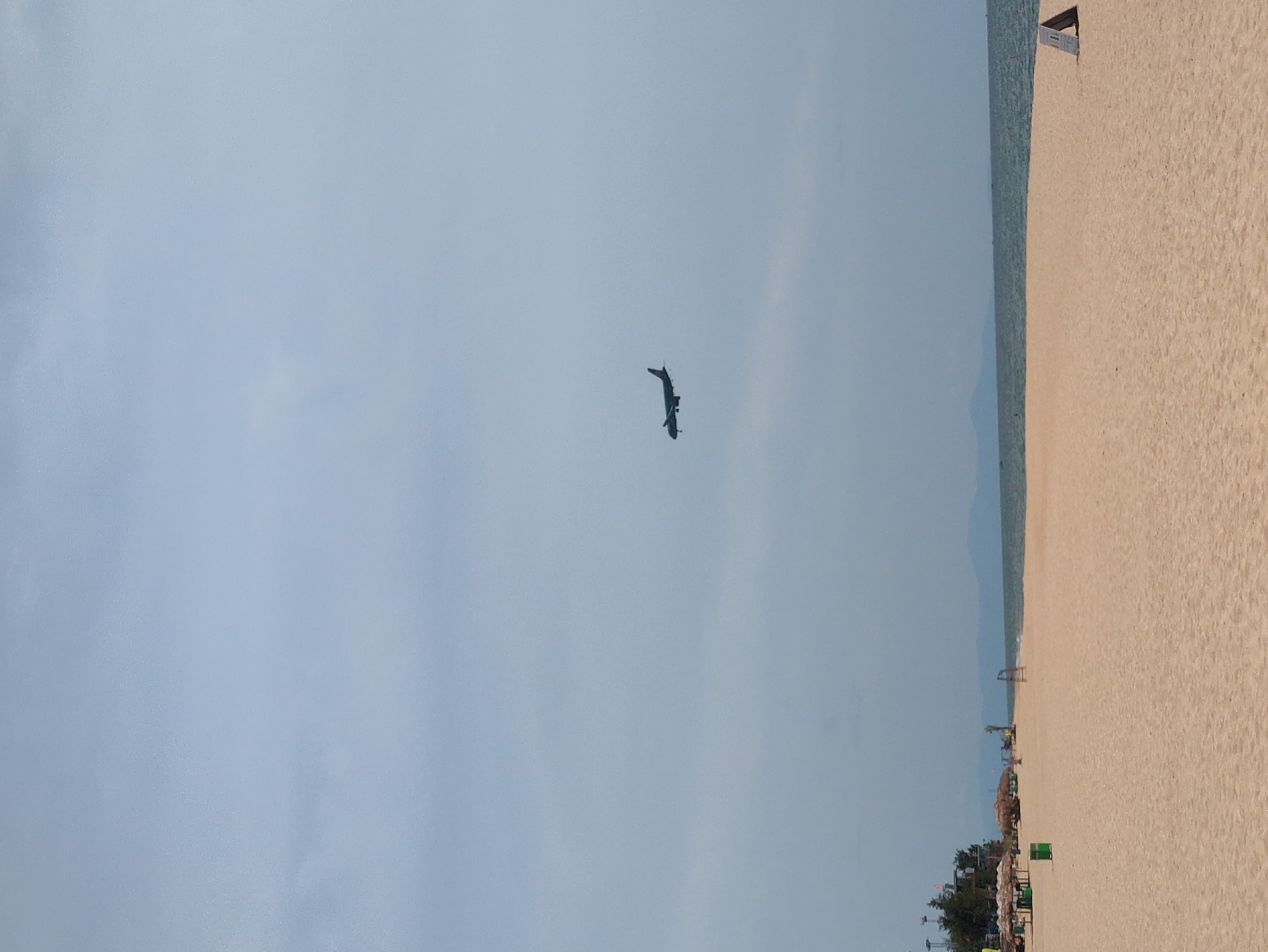 The image depicts a wide sandy beach in Đồng Hới Ward, Vietnam, under a hazy, overcast sky. In the distance, the calm, blue sea meets the horizon, with faint mountain silhouettes visible on the far shore. The sand is a light, warm beige, stretching across the foreground and midground.
Scattered on the beach are several objects. To the left, a few thatched-roof umbrellas and what appear to be green trash receptacles are visible, along with some trees and structures that suggest a resort or public area. Further to the right, a simple goalpost structure stands on the sand. In the far right corner, a small, triangular sign rests on the beach.
The most prominent subject in the sky is a silhouetted airplane in mid-flight, positioned in the upper right quadrant of the image, appearing to be on approach or departure. There are no people visible on the beach. The overall impression is one of a quiet, somewhat desolate beach day.