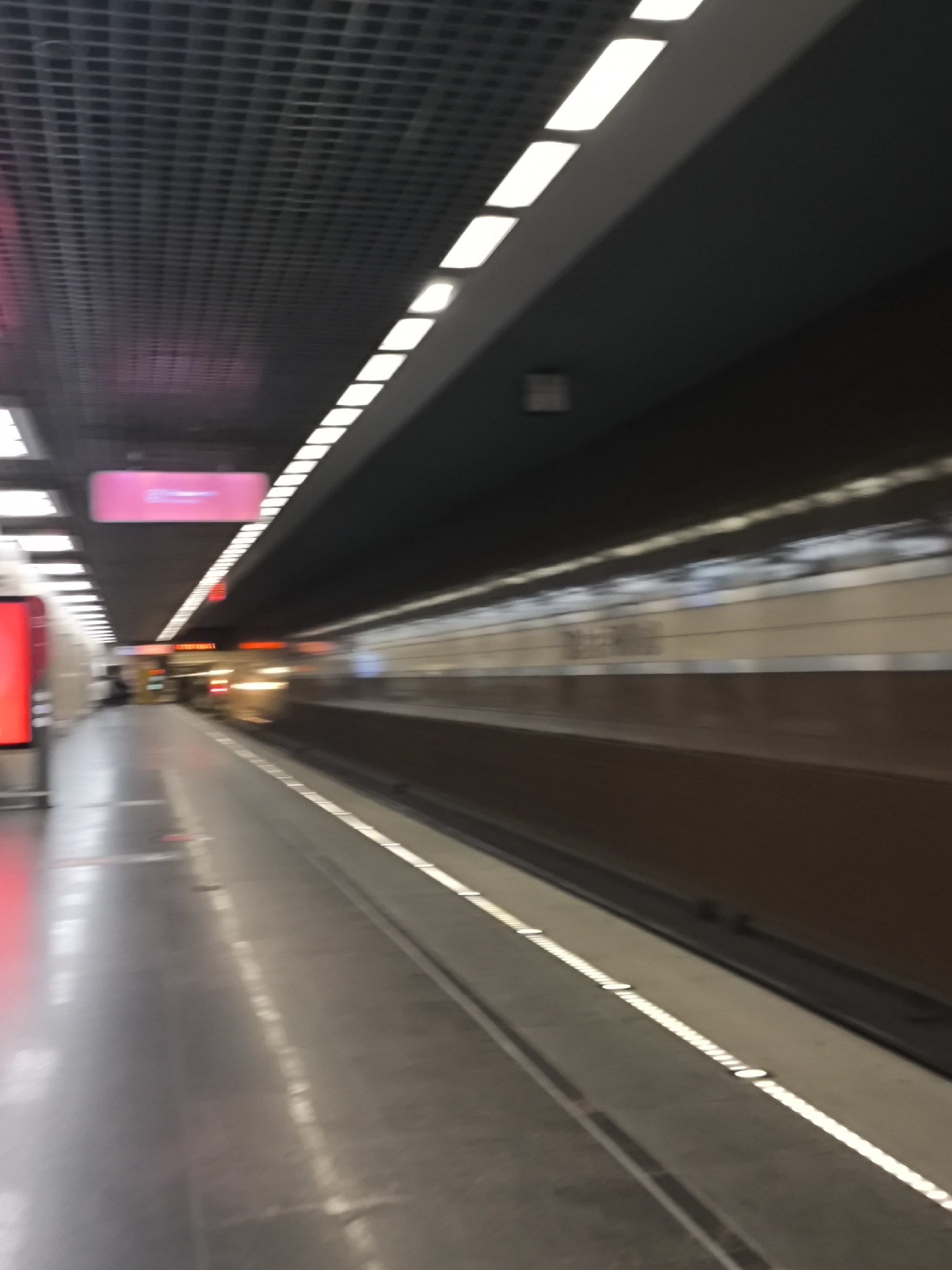 The image shows a subway platform in Moscow, Russia. A train is arriving or departing, indicated by the blurred motion. The platform is lined with white lights along the edge. An illuminated sign hangs overhead. The ceiling has a grid pattern with recessed lighting. A red advertisement or informational display is visible on the left. There are few people visible on the platform.