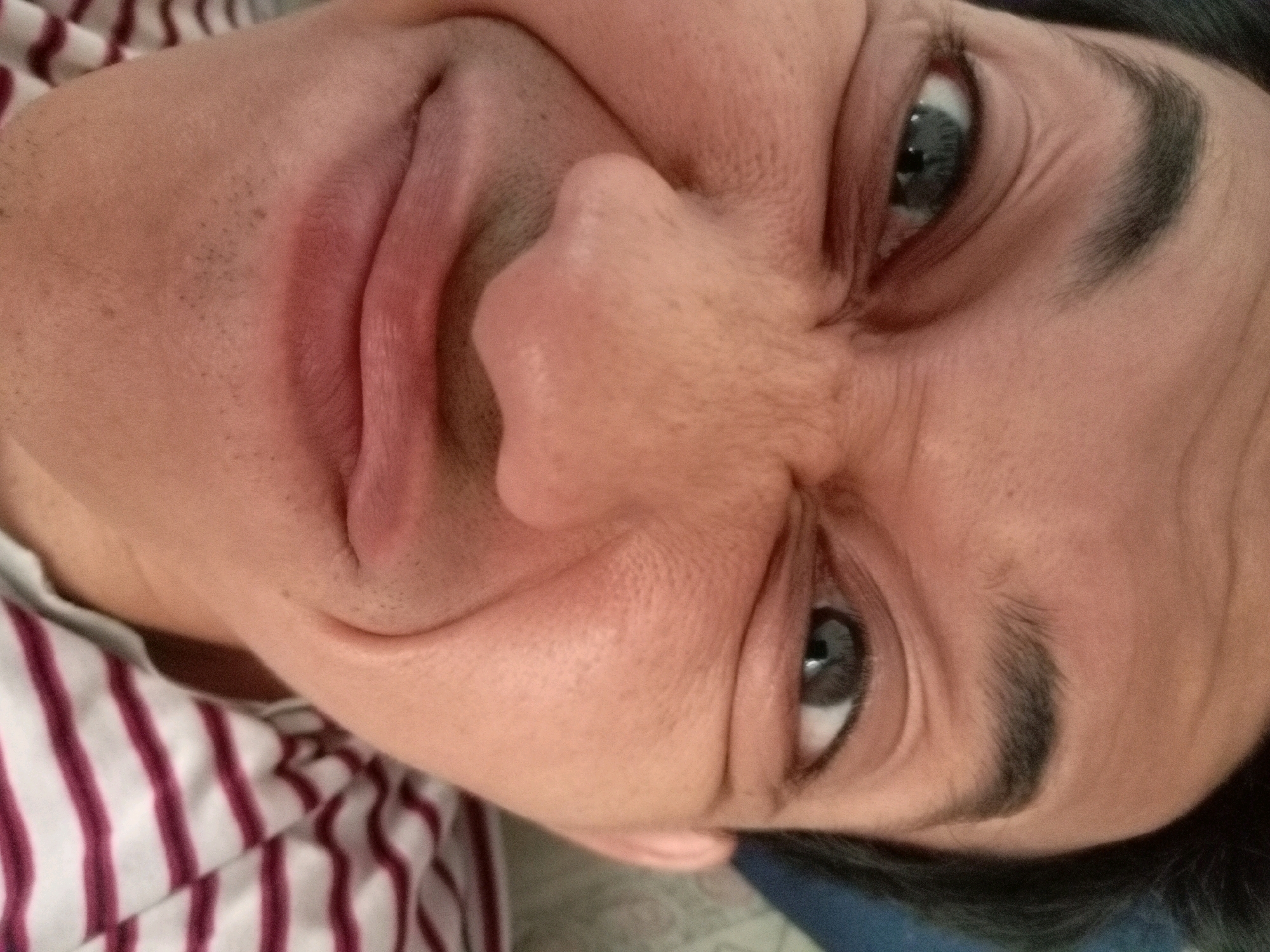 A close-up, low-angle, upside-down, portrait-style photograph shows the face of a person wearing a red and white striped shirt. The person's skin is visible in detail, including pores and stubble. The person's left eye is open and looking forward, with a blue iris. The person's mouth is partially open, showing lips with a reddish hue. Wrinkles are visible around the eyes and mouth, suggesting a contorted expression. The background is out of focus, but appears to be a bedsheet or similar fabric. There is no discernible information to determine the location in Batam, Indonesia.