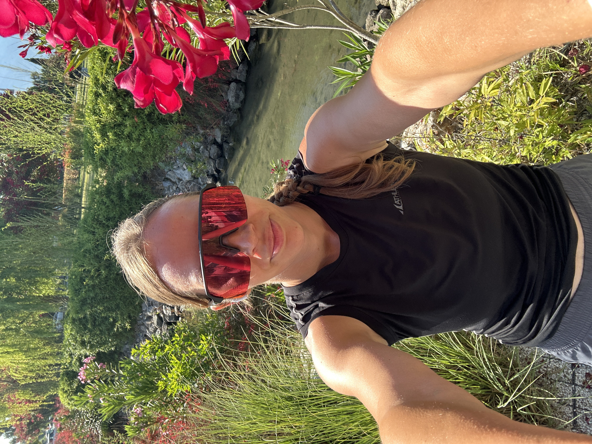The media file is a bright, sunlit selfie of a woman taken outdoors in a lush, natural environment. She is positioned slightly off-center, looking towards the camera with a subtle smile. The woman has light brown hair styled in a braid and wears large, reflective red sports sunglasses that mirror the surrounding greenery. She is dressed in a black t-shirt, which has the word "ATHLETA" visible in white text on the left chest, and dark grey shorts. Her arms are raised, indicating she is holding the camera to take the selfie.
The background is a vibrant outdoor scene, rich with diverse green foliage, bushes, and tall grasses. In the upper left and behind her head, clusters of bright red flowers, likely oleanders, hang into the frame. The mid-ground features a narrow body of clear, calm water, resembling a stream or canal, bordered by rocky banks and more dense vegetation. The scene is well-lit, indicating a clear, sunny daytime. The overall impression is one of a refreshing and scenic natural setting, consistent with a warm climate like Fethiye, Turkey.