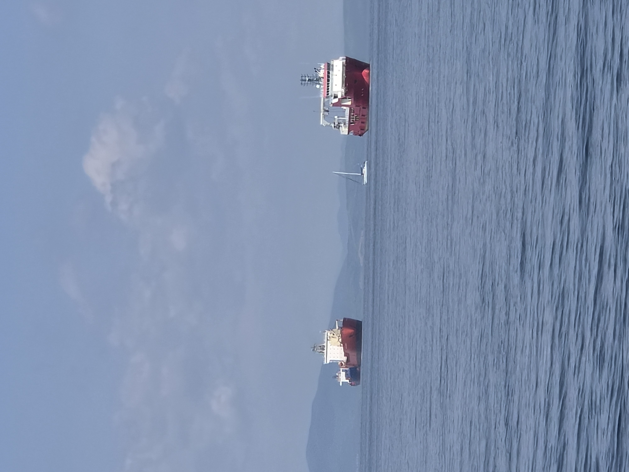 The image depicts a calm sea with several vessels. In the foreground, the water is a deep blue, with gentle ripples. In the middle ground, a large red and white ship, likely a cargo vessel or a supply ship, is positioned on the right side of the frame. To its left, a white sailboat with its sail down is visible. Further to the left and slightly behind these vessels is another large ship, predominantly red and white, appearing to be a tanker. The background features a hazy, mountainous coastline under a pale blue sky with scattered white clouds. The scene suggests a maritime setting, possibly in the waters off Vladivostok, Russia, given the presence of large ships and the mountainous terrain.