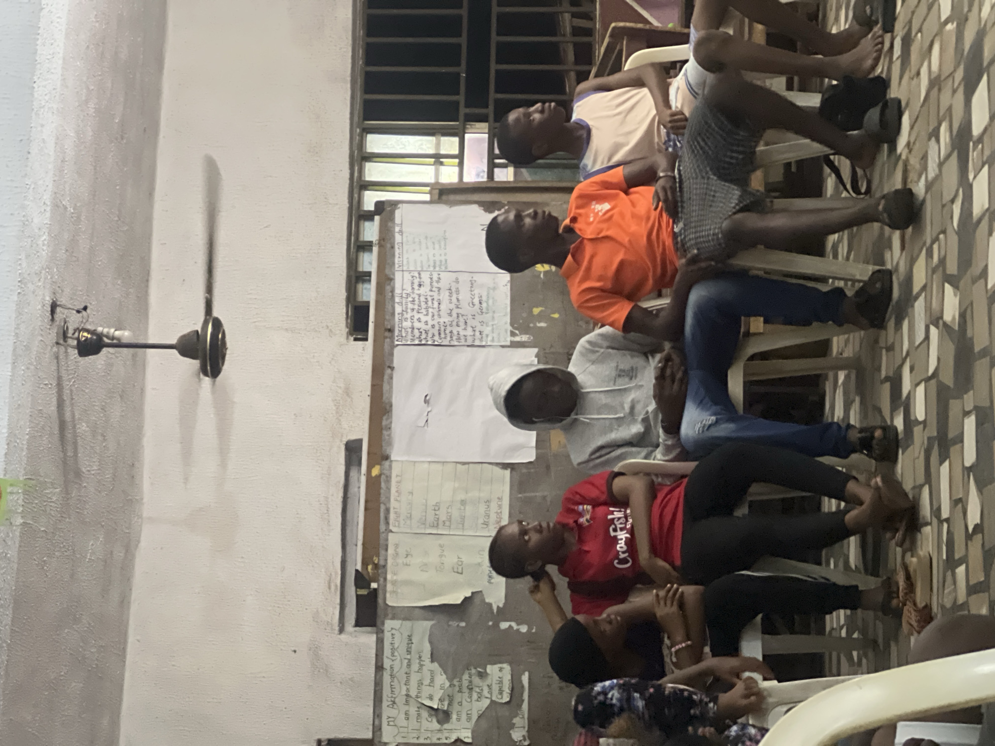 In Nkpor, Nigeria, a group of young individuals is seated in a room with a rough, textured ceiling where a ceiling fan is visible. Several whiteboards are mounted on the wall behind them, displaying handwritten notes, lists, and diagrams. The individuals, comprising both males and females, are seated on white plastic chairs, facing towards the left of the frame. They appear to be attentive, with some looking forward and others glancing to the side. The atmosphere seems relaxed, possibly during a learning session or meeting. The room's flooring is tiled in a mosaic pattern.