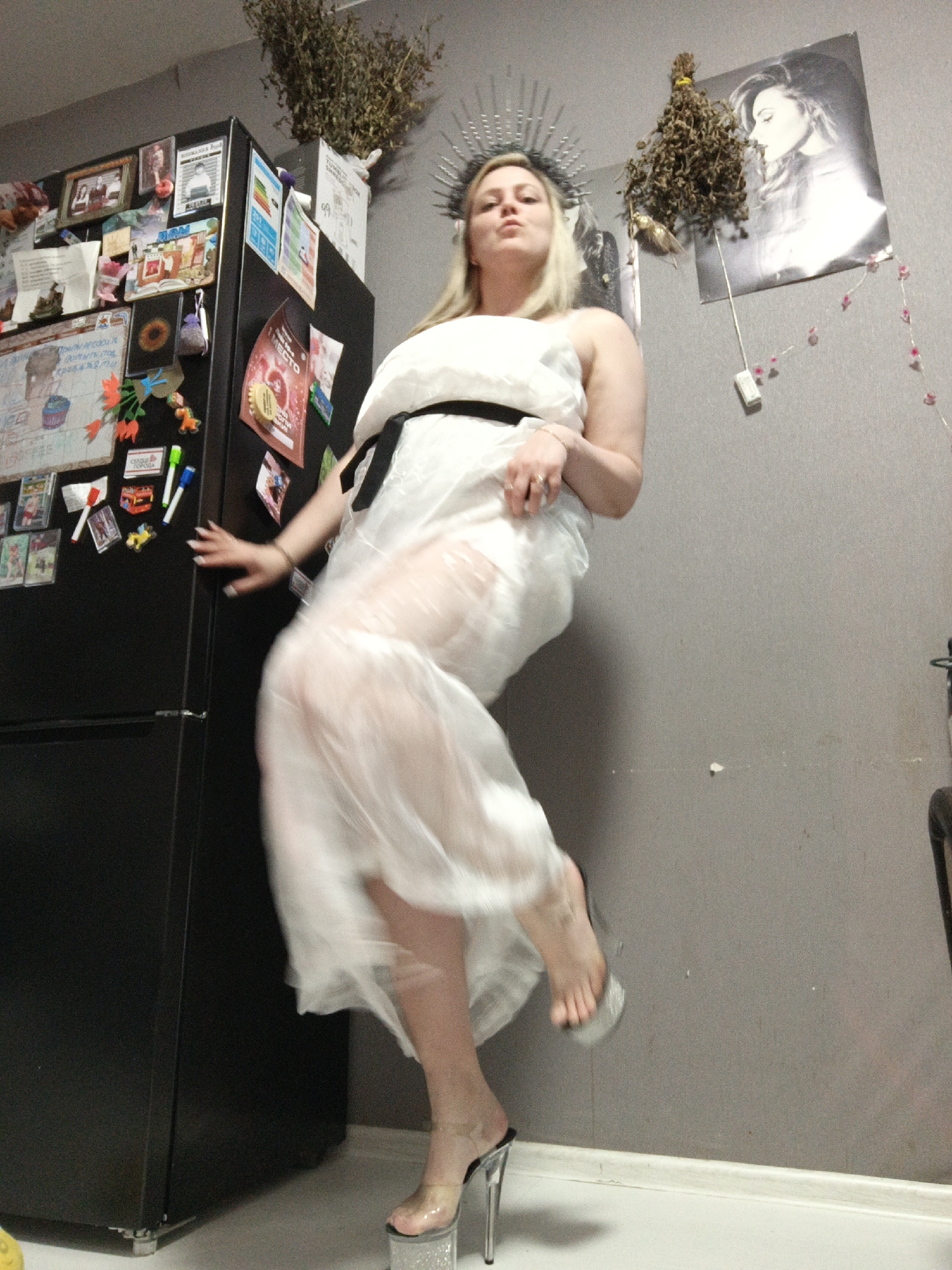 An indoor, low-angle shot captures a woman posing dramatically in what appears to be a domestic setting, possibly a kitchen or utility area. The woman, with blonde hair and a confident, slightly pursed-lips expression, wears a white, sheer, flowing dress with a black belt and a silver-spiked headpiece resembling a crown or halo. She is balancing on one foot, which is adorned with a clear platform high heel, while the other leg is lifted, causing the sheer dress to swirl around her. Her hands are somewhat blurred by motion, one resting on a refrigerator, the other near her waist.
To the left, a large, black, double-door refrigerator is visible, densely covered with an assortment of magnets, photographs, papers, and children's drawings. Visible Russian text on the refrigerator includes "1 место" (1st place) on a red flyer, "ФЕВРАЛЬ" (February) on a calendar-like item, and "С 8 МАРТА" (Happy March 8th) and "С днем рождения" (Happy Birthday) on other decorations. Another partially visible text appears to say "ЦИРК В ГОРОДЕ" (Circus in the city).
The background consists of a plain gray wall. Two bundles of dried herbs or plants hang on the wall, one above the refrigerator and another to the right. Between these bundles, a black and white poster of a woman (possibly a celebrity) is affixed to the wall. A string of pink, flower-shaped decorative lights or beads descends from the wall, connecting to a white power outlet. The floor is white, with a dark baseboard running along the wall. The scene is brightly lit by interior lighting, and the woman's pose conveys a sense of playfulness or theatricality.